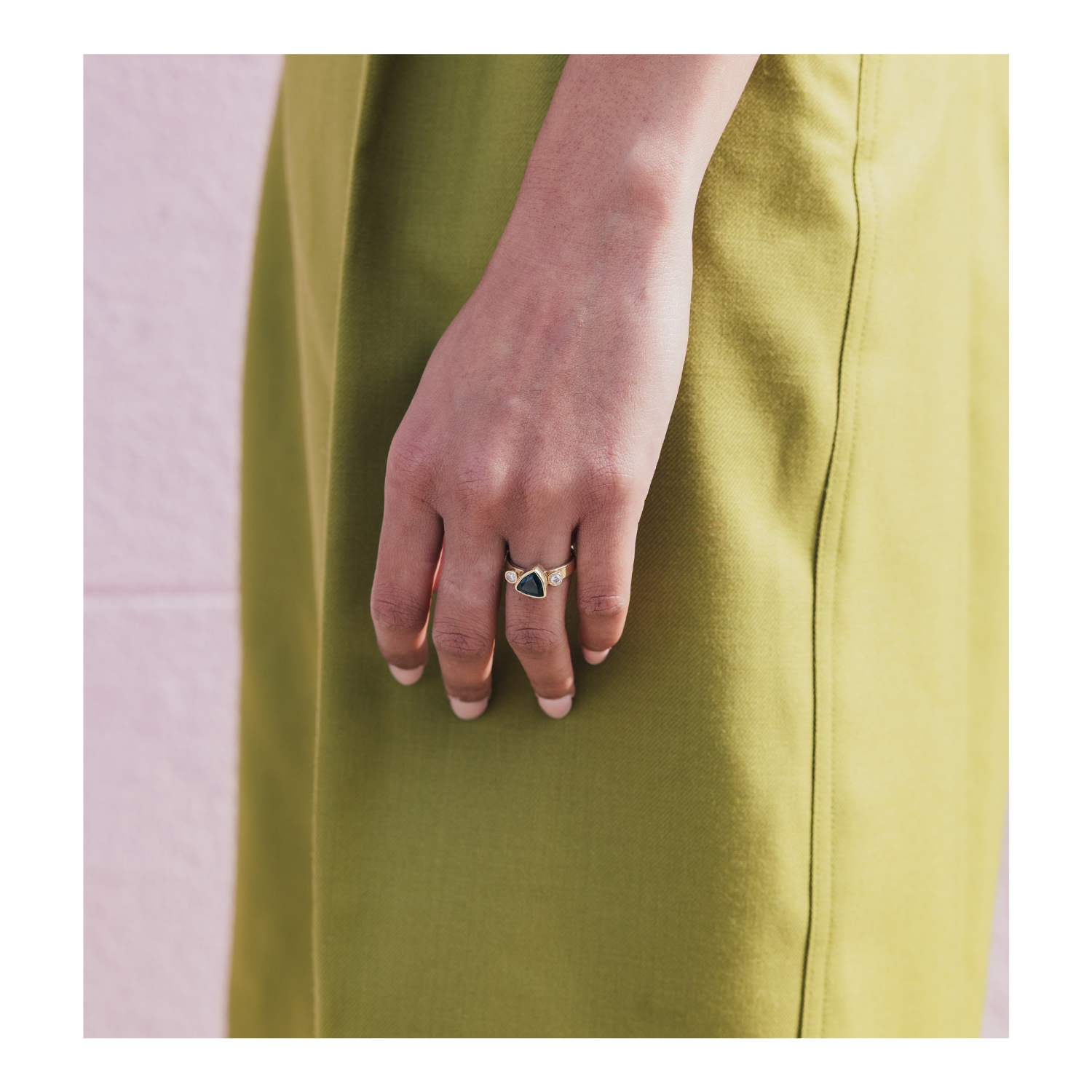 A close-up of a person's hand wearing a gold ring with a dark triangular stone, set against a solid olive green dress.