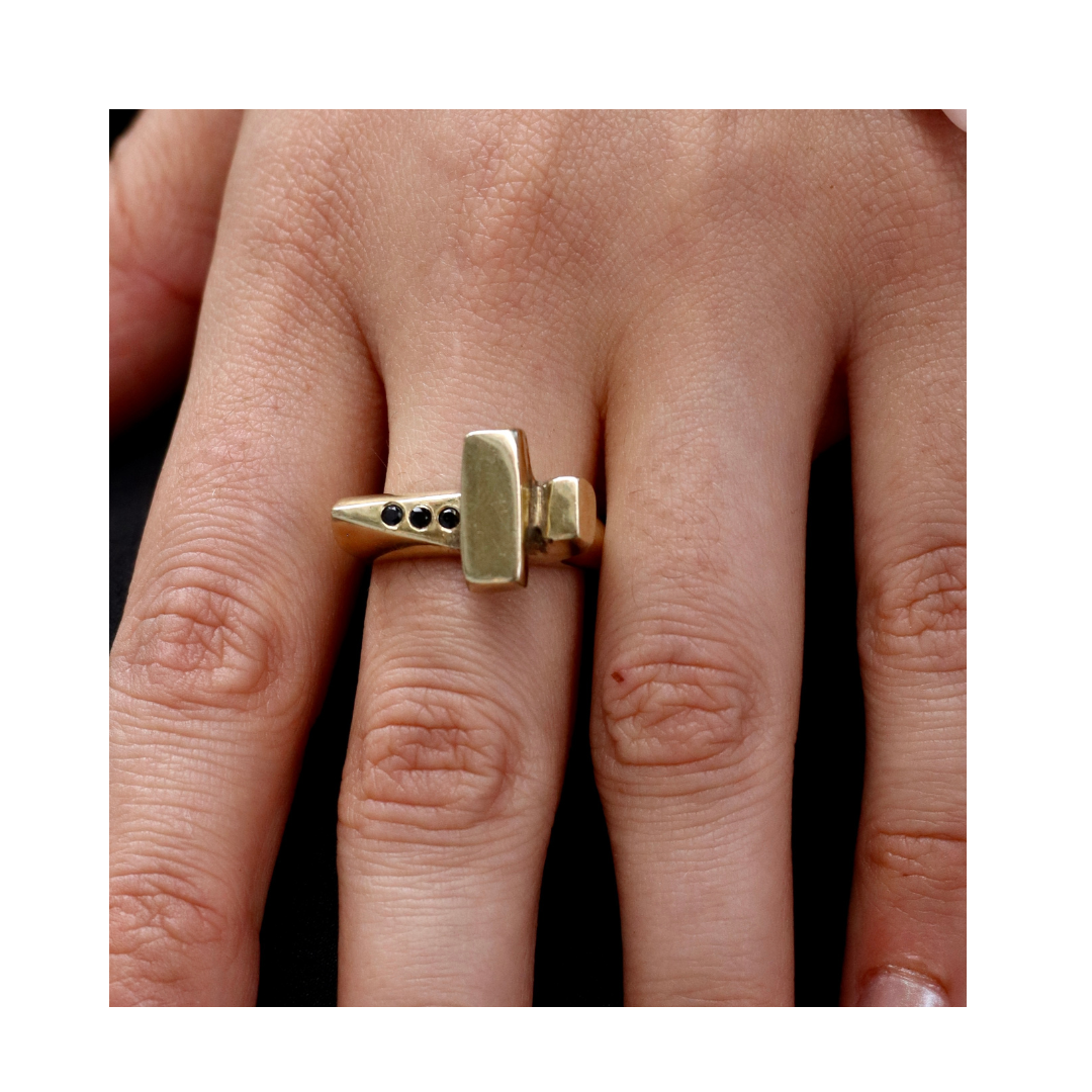 A close-up of a hand wearing the Totem Ring Brass with Black Spinel by Madeleine Sculptural Jewellery, featuring a gold recycled brass band shaped like a hammer and set with small black stones against a simple background.