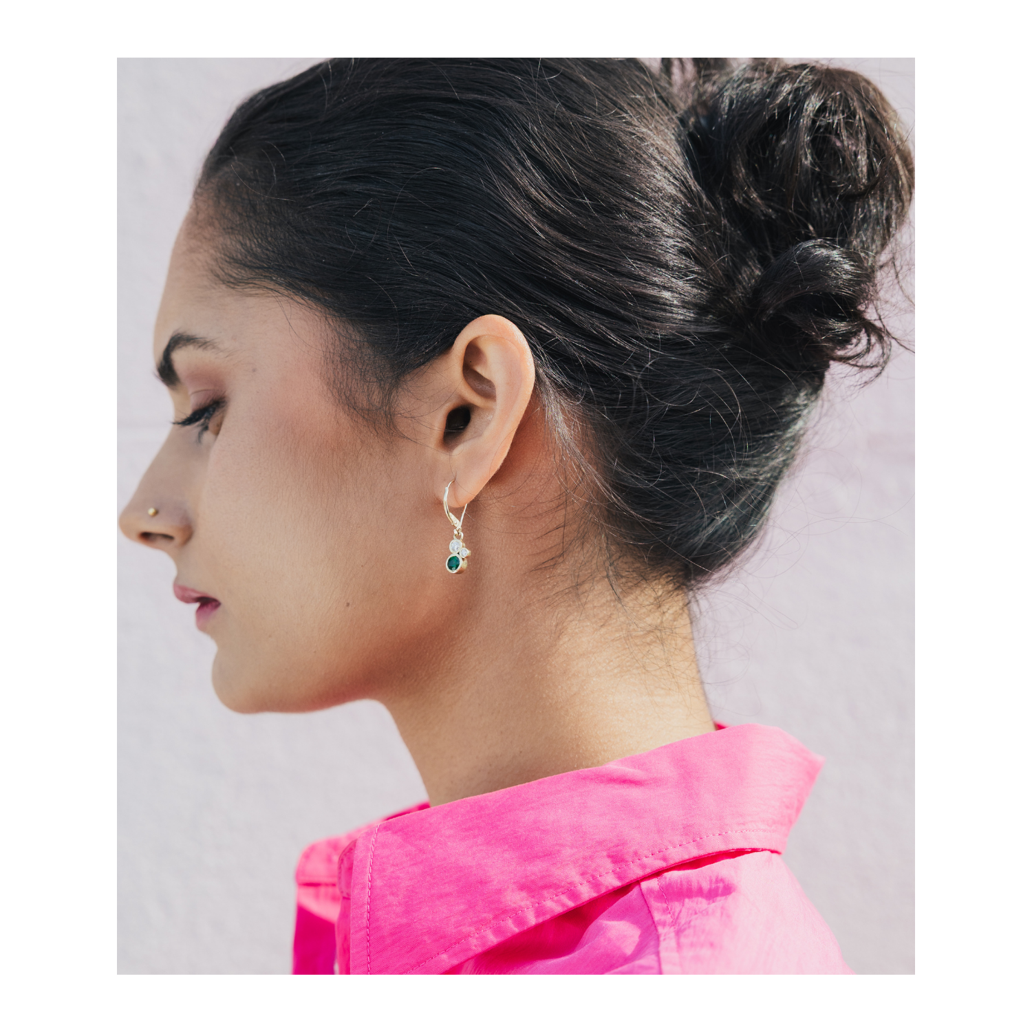 A woman in profile wearing a small silver dangle earring with a green gemstone, paired with a bright pink shirt against a light gray background.