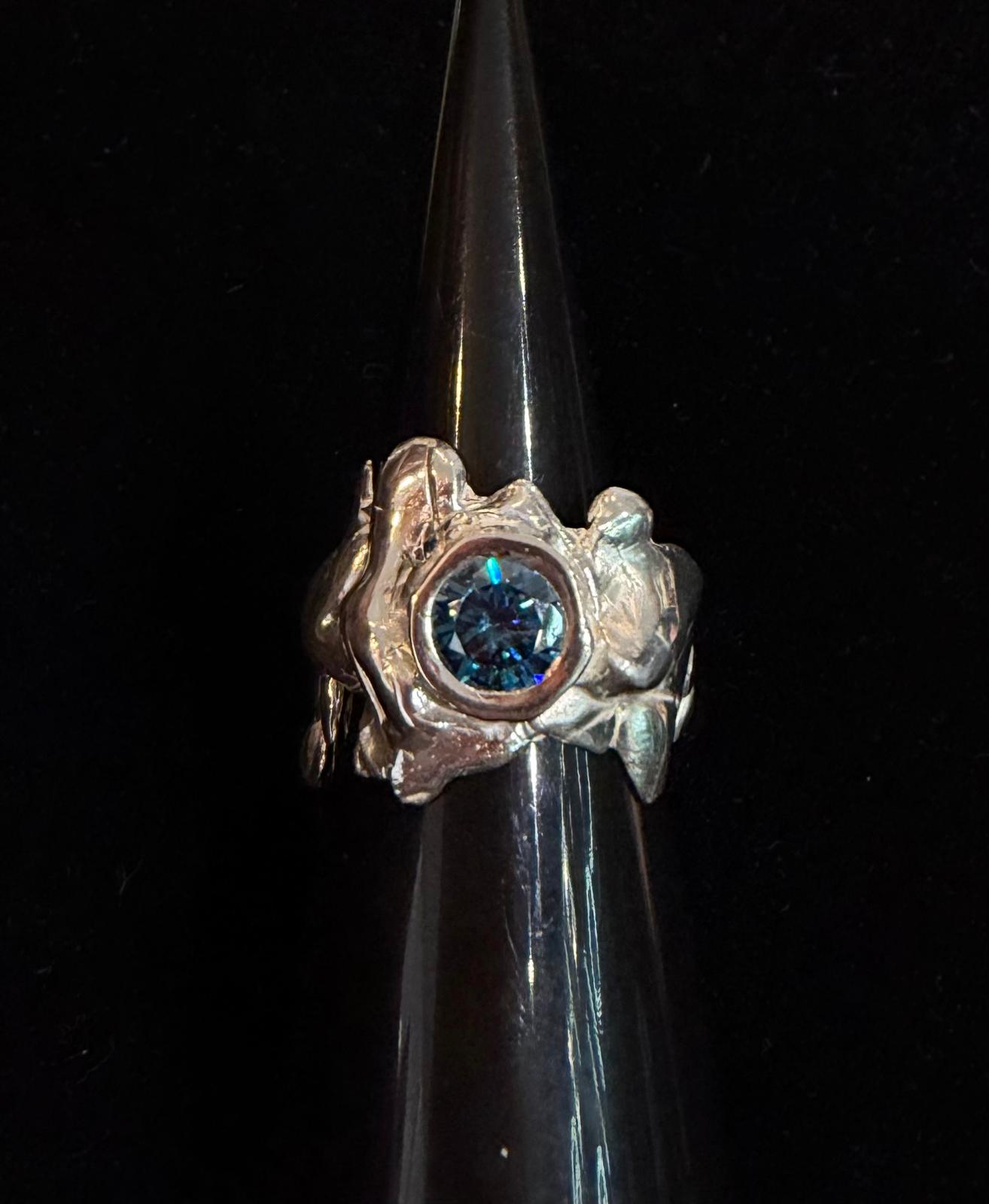 The Solstice Ring with Large Moissanite by Madeleine Sculptural Jewellery is a handcrafted silver ring with an organic, textured design, featuring a round moissanite center stone, displayed on a black glossy stand against a dark background.