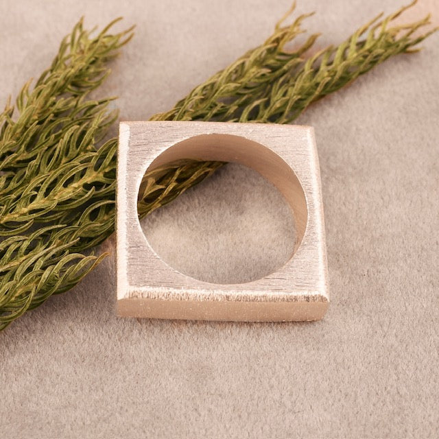 The William Silver Ring by Gallardo & Blaine Designs, featuring a square shape and brushed 925 sterling silver finish, is displayed on a beige surface beside sprigs of green plants.