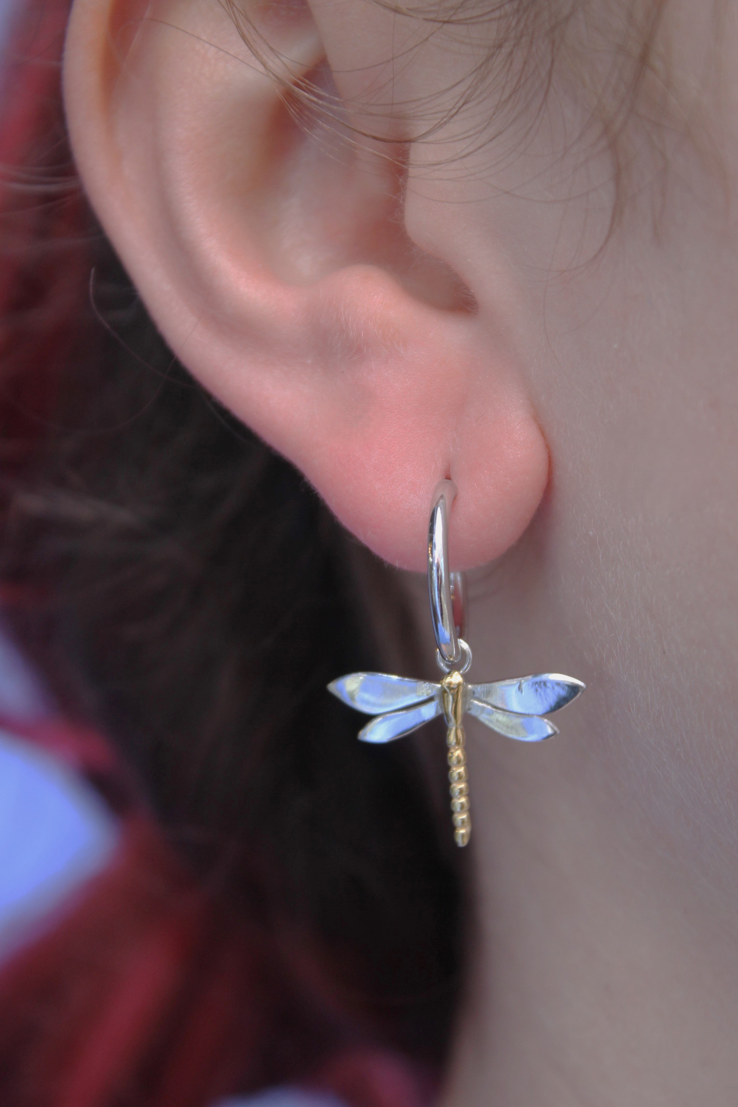 A close-up shows a light-skinned person with reddish-brown hair wearing Watermelon Tropical’s Wildlife Dragonfly Dangle Earrings in silver, featuring a dragonfly charm with light blue and gold accents. Lightweight and eye-catching.