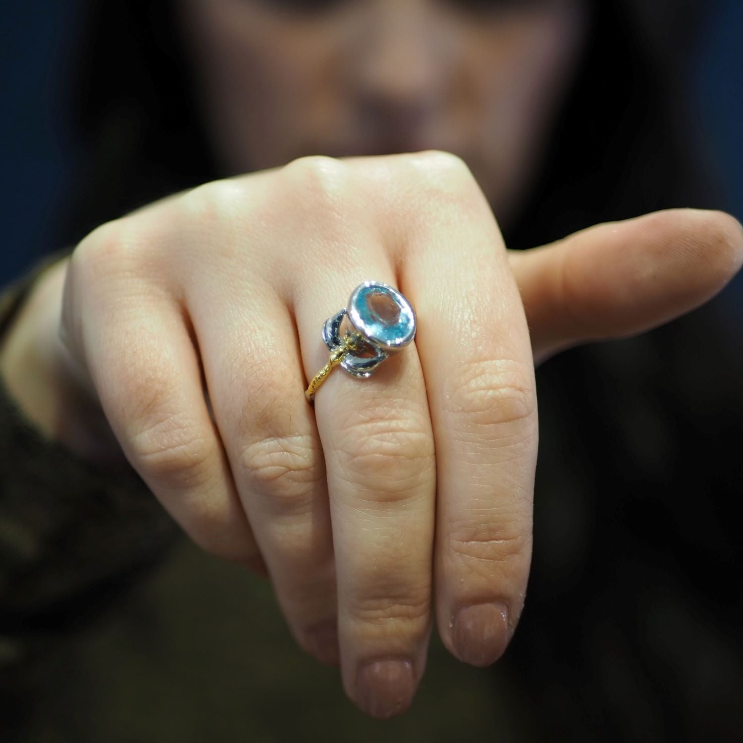 A person wearing a delicate and dainty Gallardo & Blaine Designs Daydream Ring in various gemstones with a large blue gemstone on their finger. The hand is shown up close, with the fingers partially curled and extending towards the camera, making the time