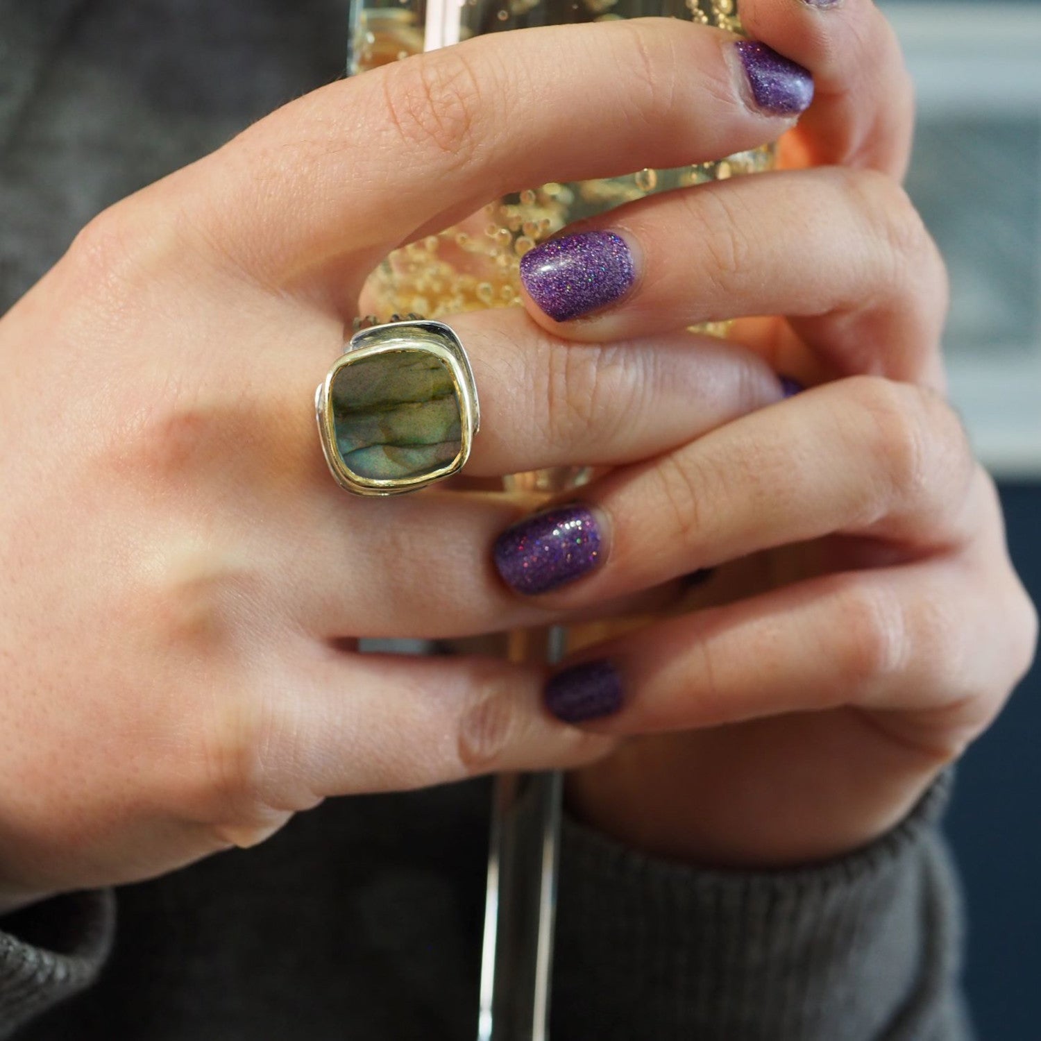 A person holds a sparkling glass with golden liquid, displaying their Adjustable Art Deco Ring in Silver Gold by Gallardo & Blaine Designs, featuring a large square gemstone. Their nails shine with purple glitter polish against an elegantly blurred backgr