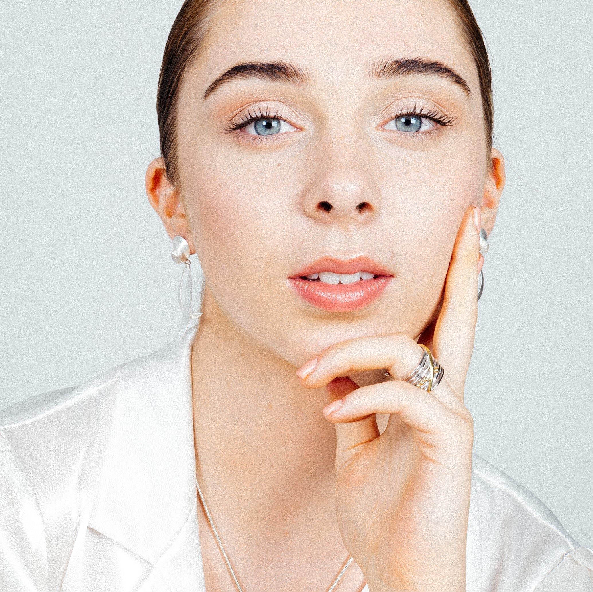 A young woman with fair skin, blue eyes, and brown hair pulled back is gazing into the camera. She is wearing a white top, gold vermeil earrings, and a Gallardo & Blaine Designs Bamboo Ring in Sterling Silver & Gold Vermeil on her index finger. Her hand r