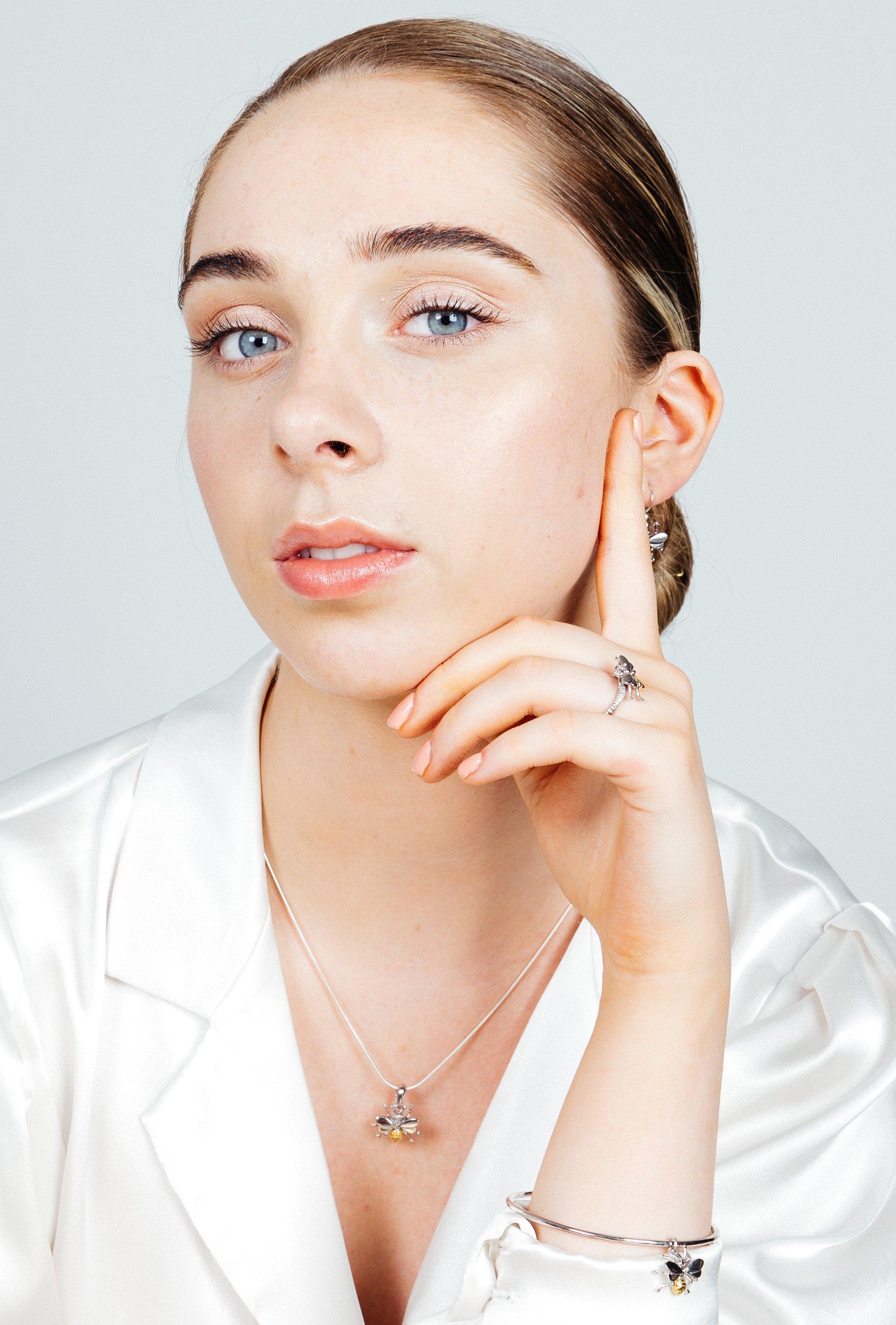 A woman with blue eyes and light skin wears a white satin top and elegant silver and gold jewelry, featuring the Wildlife Bee Bangle by Watermelon Tropical. Her slicked-back hair frames her face as she gazes at the camera, hand resting on her cheek.