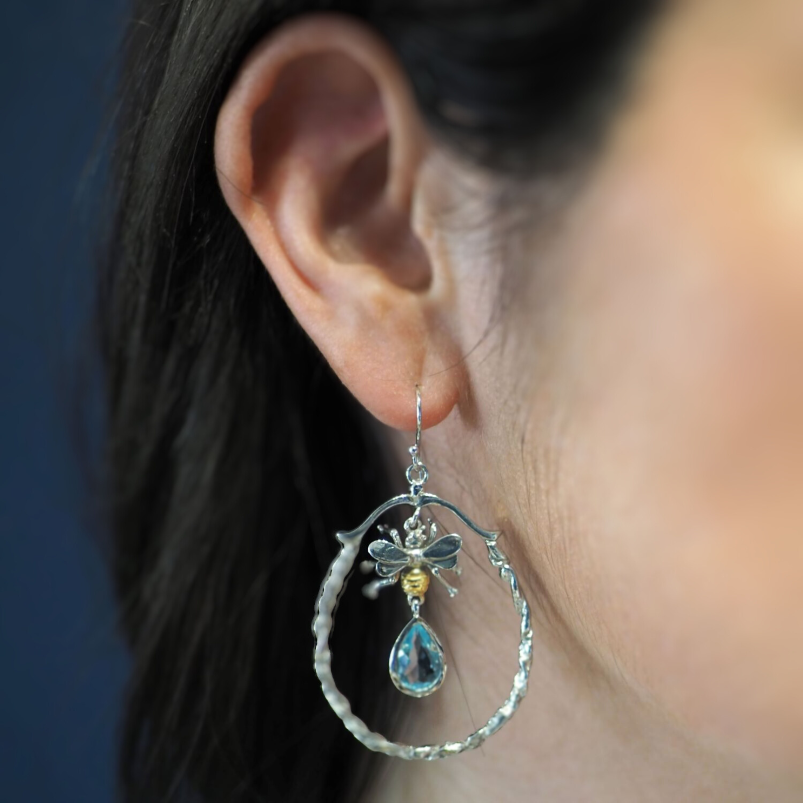 A close-up of a person’s ear wearing Watermelon Tropical Wildlife Large Bee Hoop Drops, featuring a bee at the top and a blue teardrop gemstone inside textured silver hoops.
