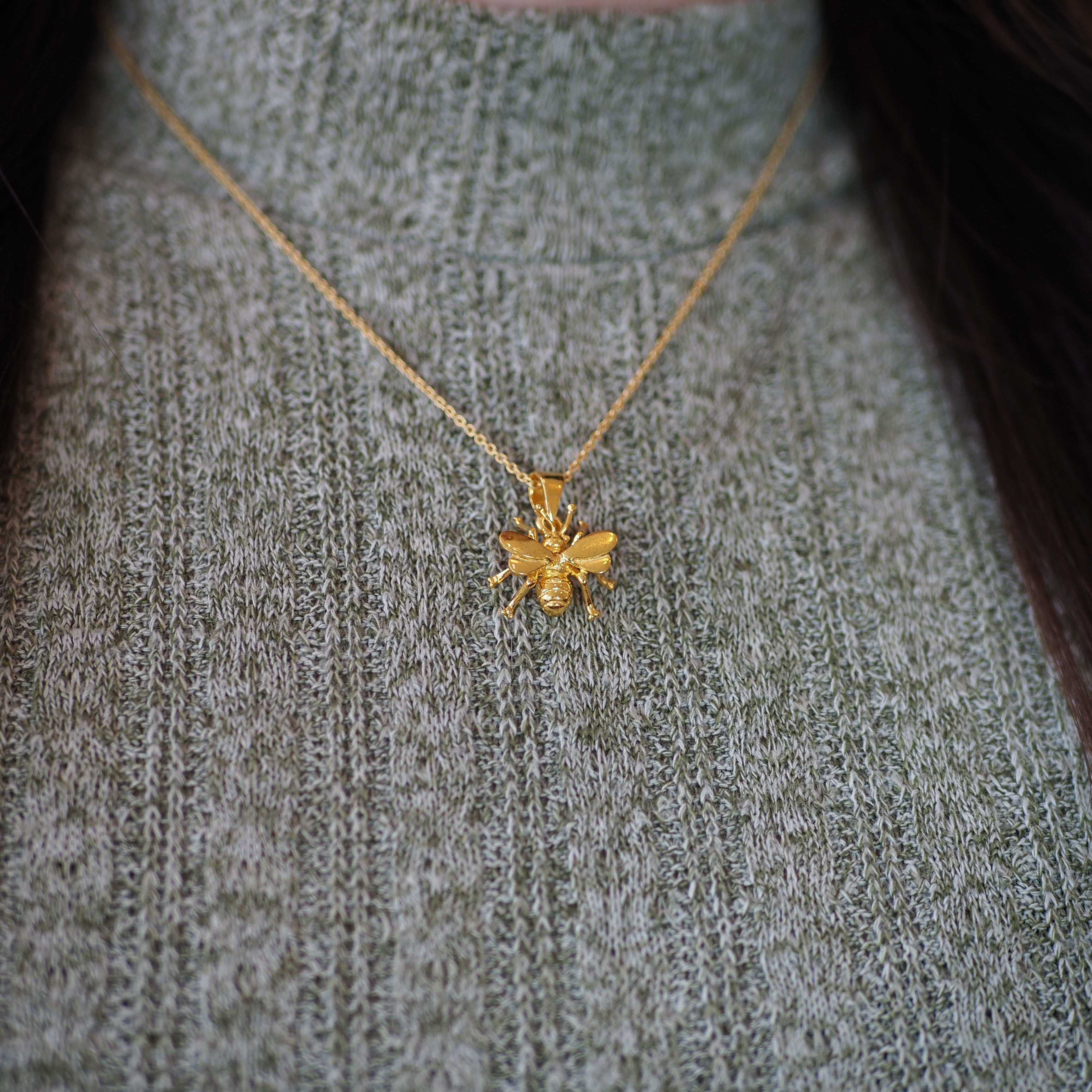 The Watermelon Tropical Wildlife Pendant - Small Bee With Chain is displayed on a light grey-green ribbed turtleneck, featuring gold-detailed wings that elegantly hang from a matching gold chain necklace.