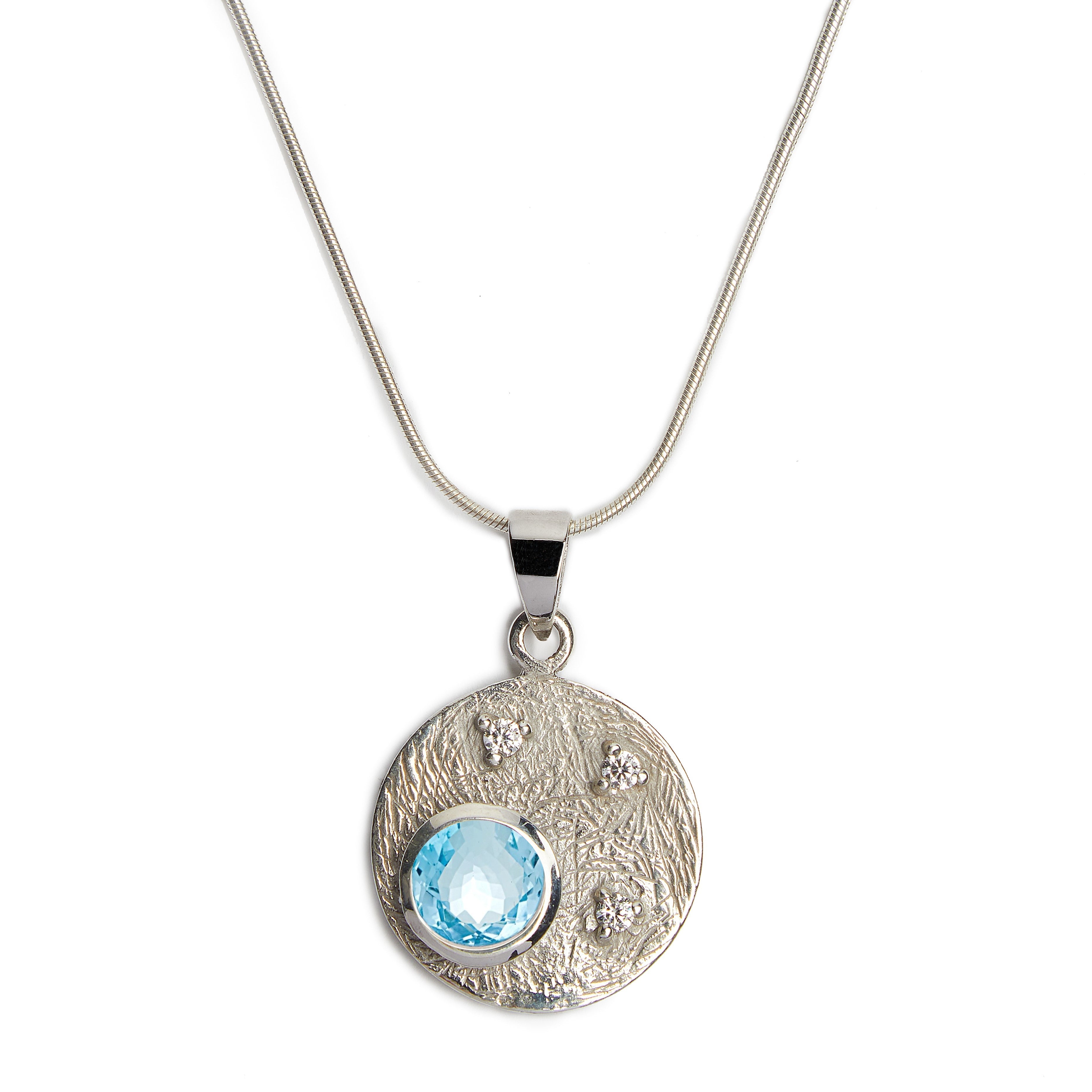 Introducing the Celeste Necklace in Silver & various gemstones by Gallardo & Blaine Designs, a sterling silver piece featuring a round pendant. The pendant showcases a textured design with small embedded crystals and a larger central blue cubic zirconia g