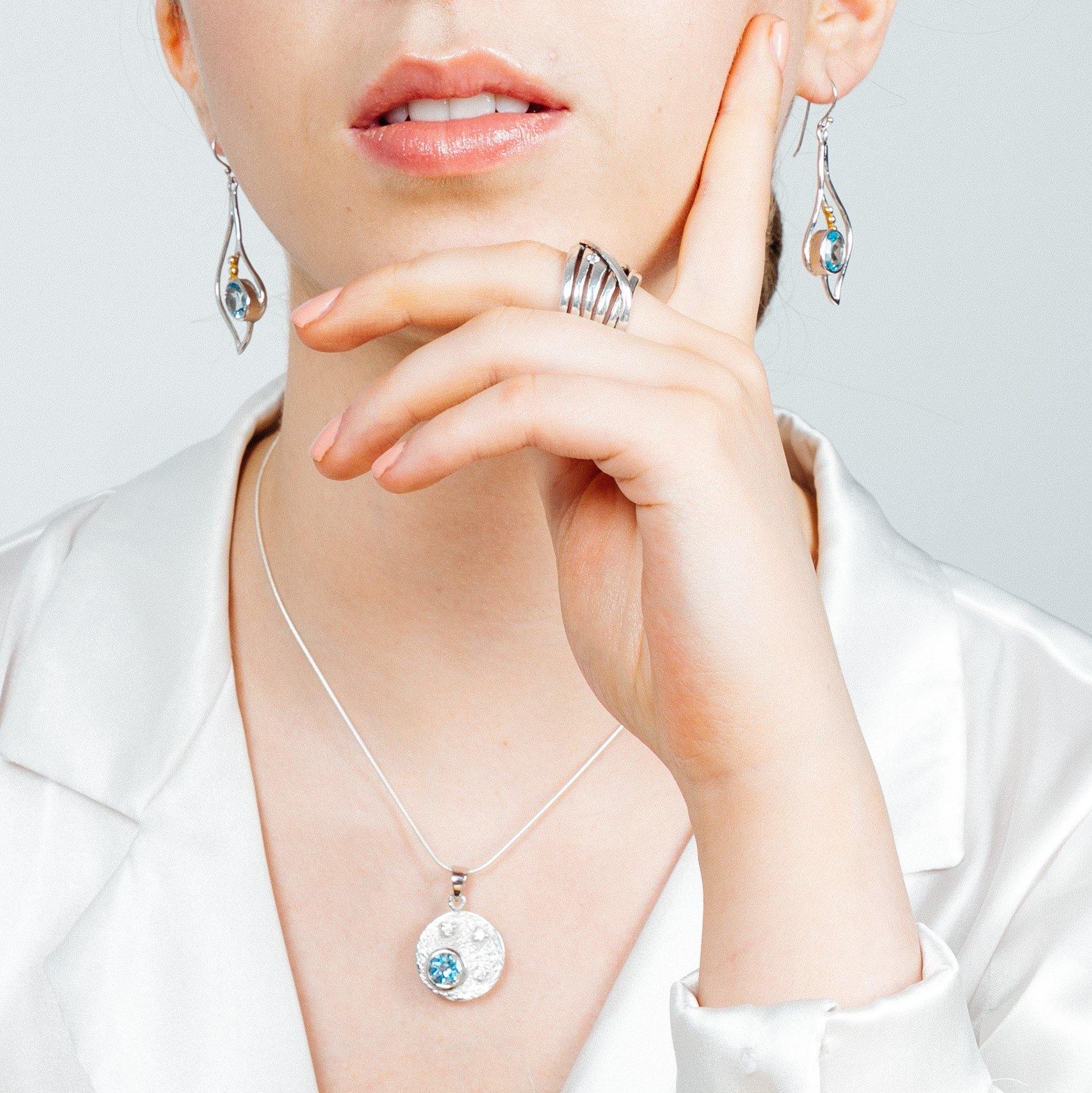 A person wearing elegant jewelry, including dangling earrings with blue stones, a **Gallardo & Blaine Designs Celeste Necklace in Gold & various gemstones**, and a stylish multi-band ring. The person is dressed in a white satin blouse and holds their hand