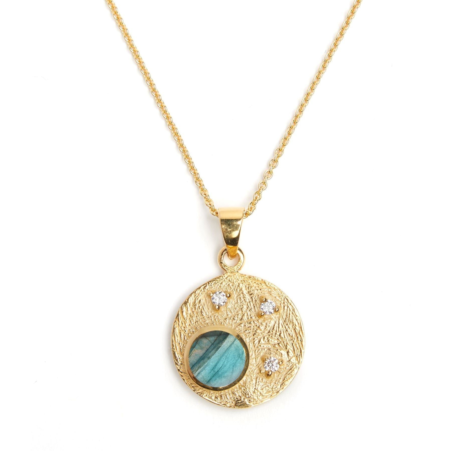 A Gallardo & Blaine Designs Celeste Necklace in Gold & various gemstones with a textured circular pendant featuring a blue-green gemstone in the center, accented by three small white cubic zirconia stones arranged in a triangular pattern. The stunning gem