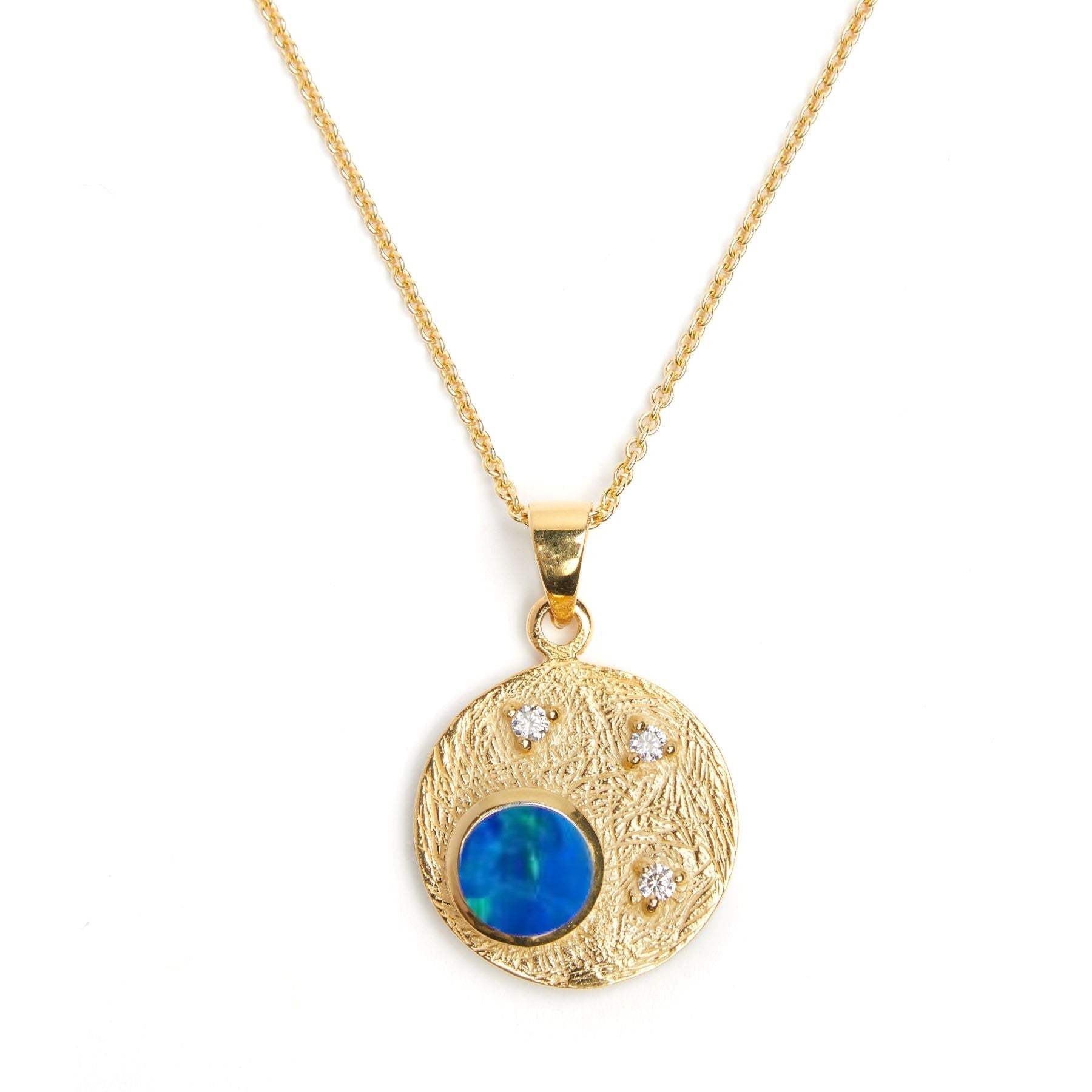 A Gallardo & Blaine Designs Celeste Necklace in Gold & various gemstones with a textured circular gemstone pendant featuring a round blue gemstone and three small clear cubic zirconia stones is shown on a white background. The pendant hangs from a delicat