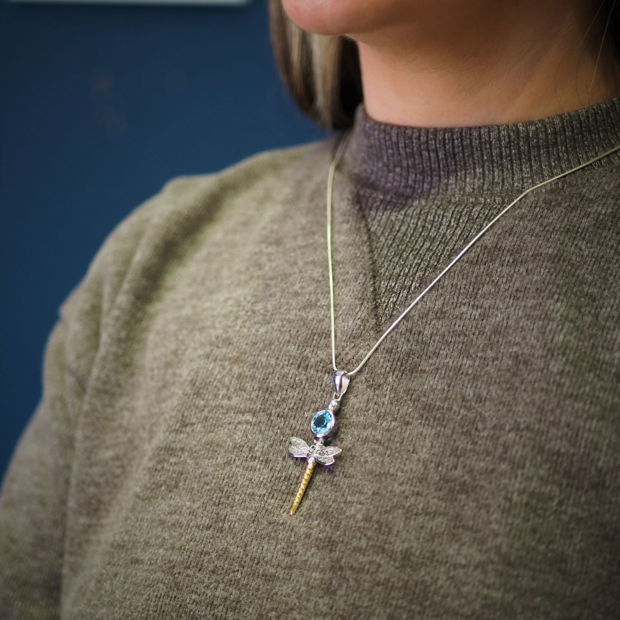 A person wearing a green sweater is shown from the shoulders up. They're adorned with a Damsel Pendant in various gemstones, a sterling silver necklace shaped like a sword or dagger, showcasing a blue gemstone in the hilt and small wings near the blade by