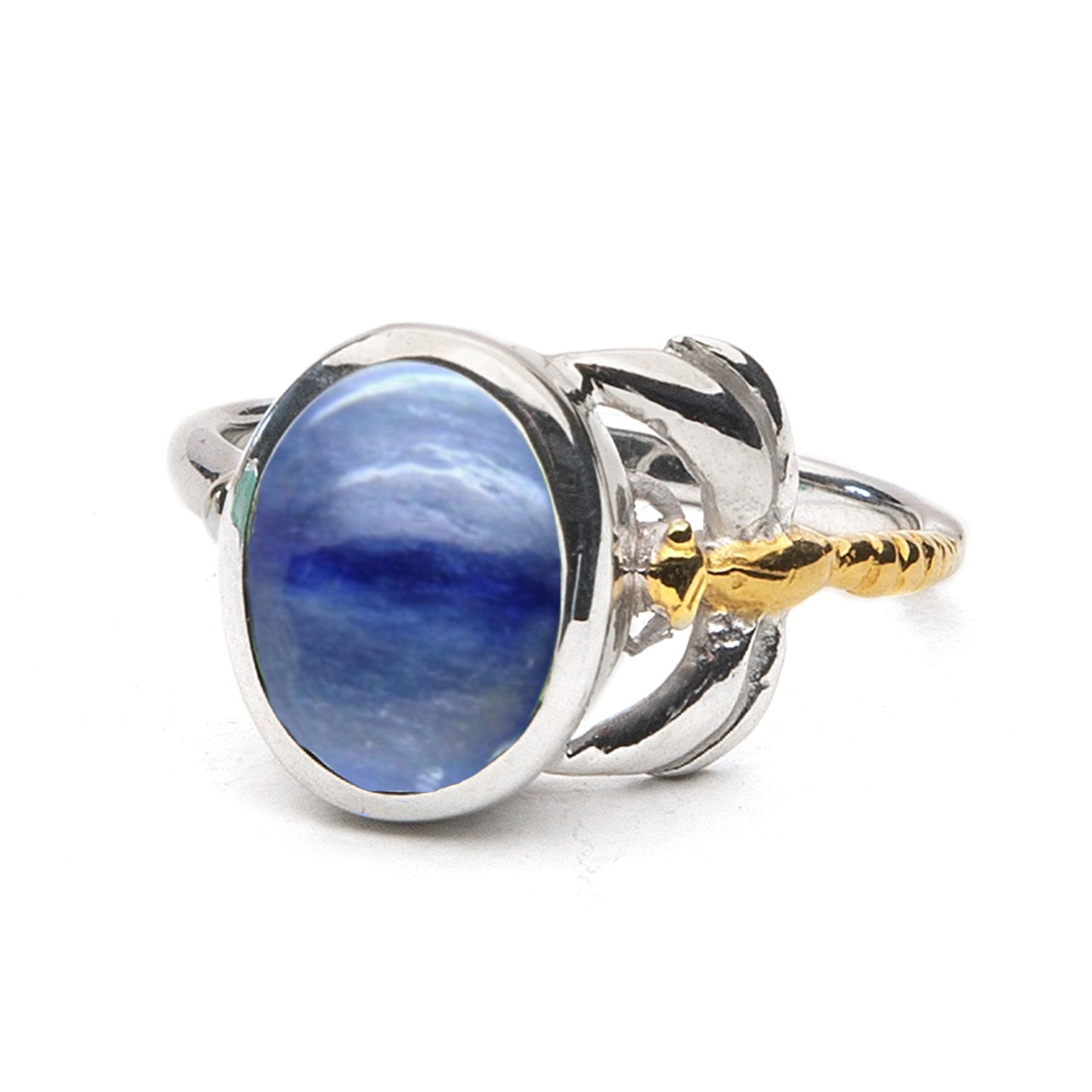 A silver ring featuring a large oval blue gemstone set in the center. The stone is held by intricate silver prongs with decorative yellow gold accents on either side of the band, creating an elegant and luxurious design. This timeless piece is perfect for