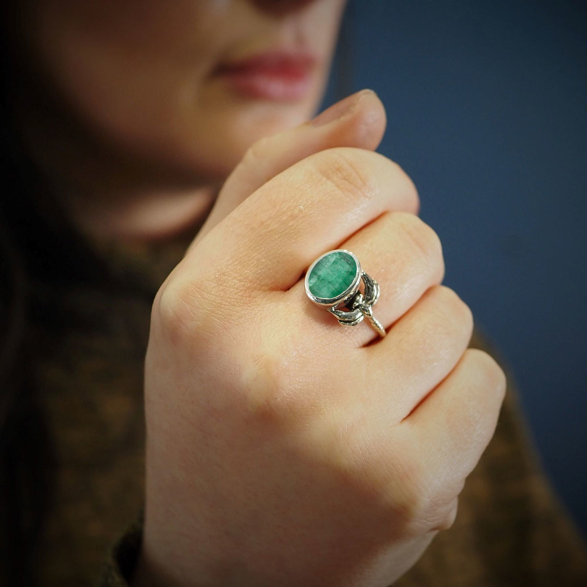 A person holds their hand near their face, wearing a Gallardo & Blaine Designs Daydream Ring in a large green gemstone set in sterling silver gold plate, with a blurred blue background.