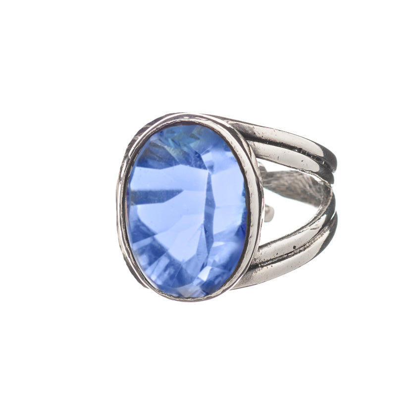 A close-up image of the Dewberry Ring in various gemstones featuring a large oval-shaped blue gemstone by Gallardo & Blaine Designs. The sterling silver band is textured and splits into two near the faceted stone. Set against a white background, the ring'