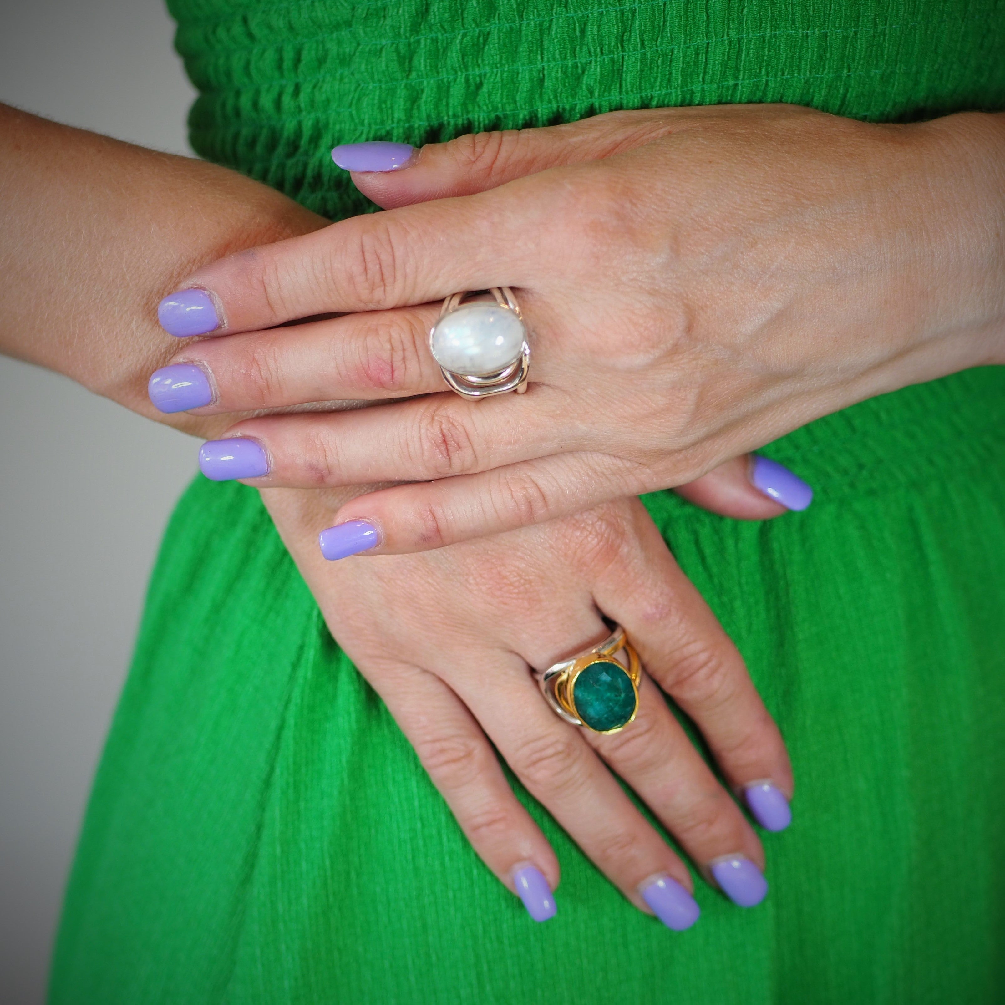 A person with light skin wearing a green dress has their hands crossed. They have lavender nail polish and are wearing two chunky statement rings: one Dewberry Ring in various gemstones by Gallardo & Blaine Designs with a large white stone on the middle f