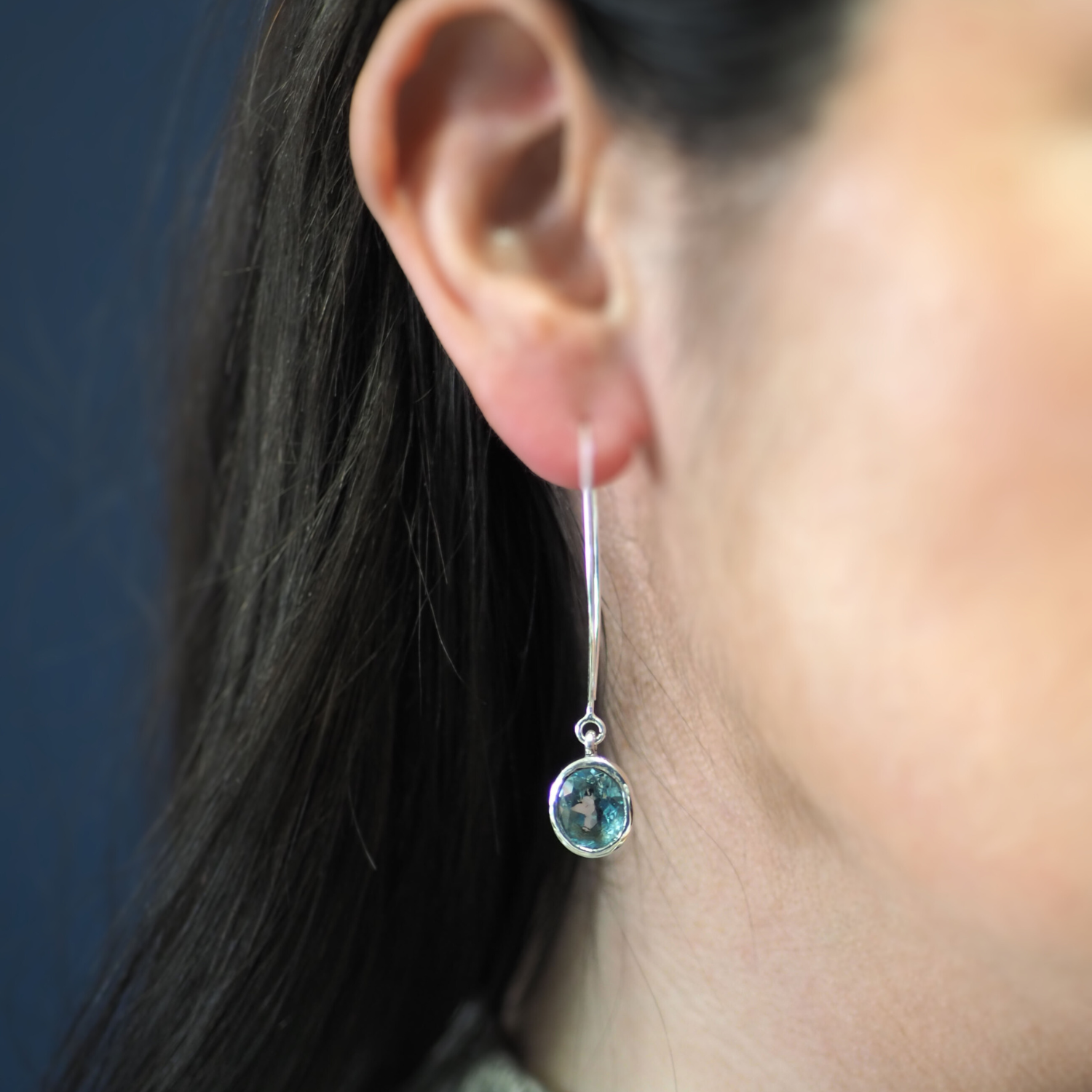 Close-up of a person wearing a long, elegant hoop design earring featuring a round, greenish-blue gemstone. The person's ear and part of their dark hair are visible against a blurred dark background, highlighting the sterling silver elegance of the Dew Dr