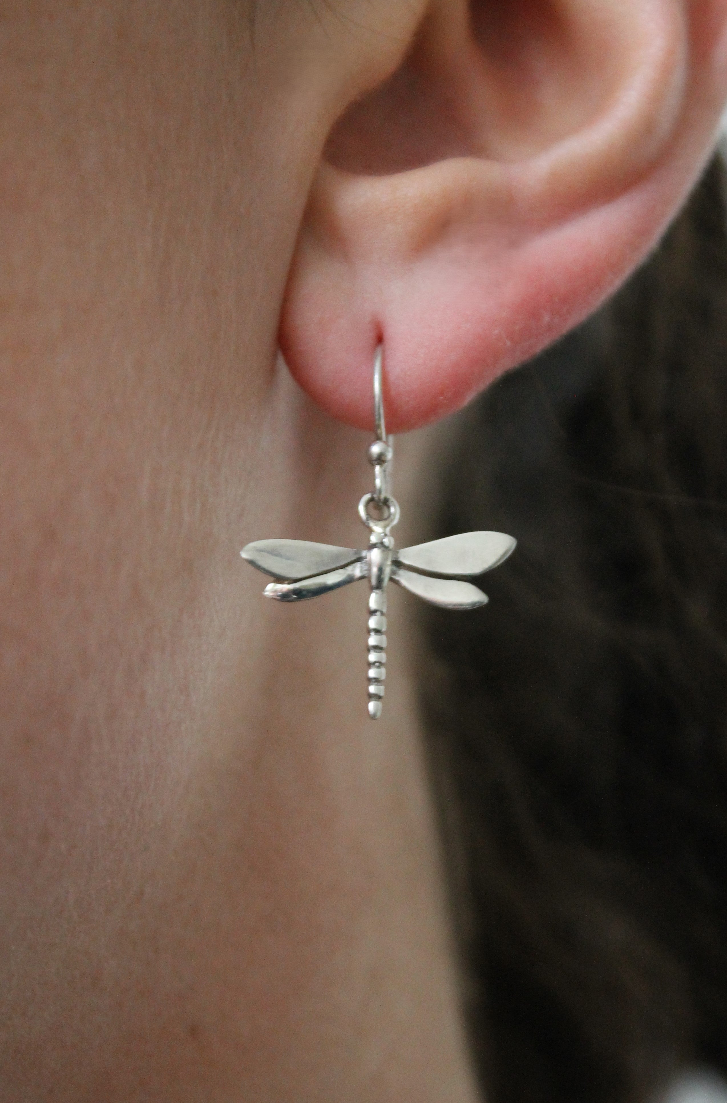 A close-up of an ear wearing Watermelon Tropical's Wildlife Dragonfly Dangle Earrings in silver, highlighting the delicate wings and body. These lightweight earrings elegantly hang from a hook and are available in silver or gold.