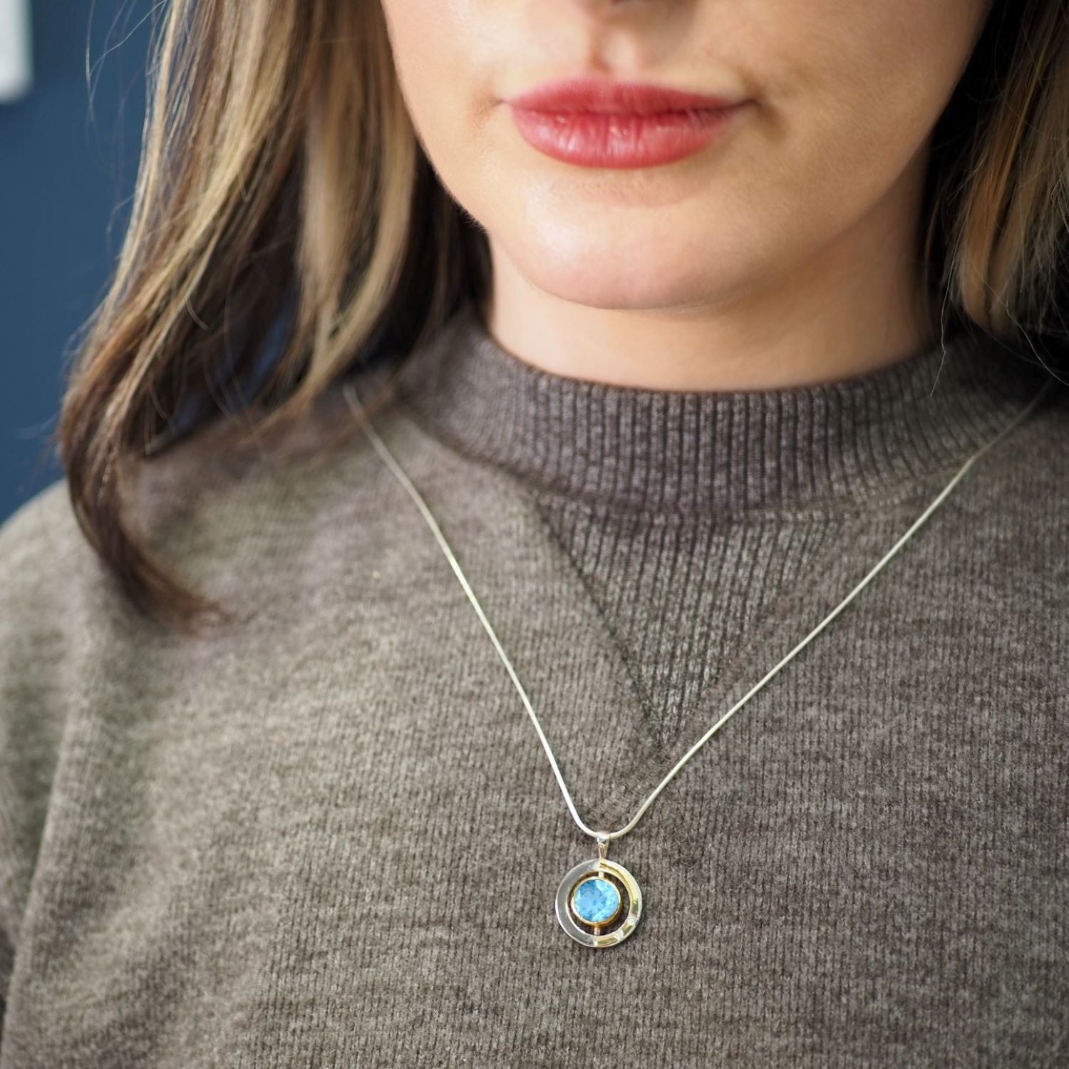 A close-up of a person wearing a brown sweater and an Elodie Pendant in silver gold & various gemstones from Gallardo & Blaine Designs. The sterling silver pendant features a blue gemstone in the center. The person has light-colored hair with darker strea