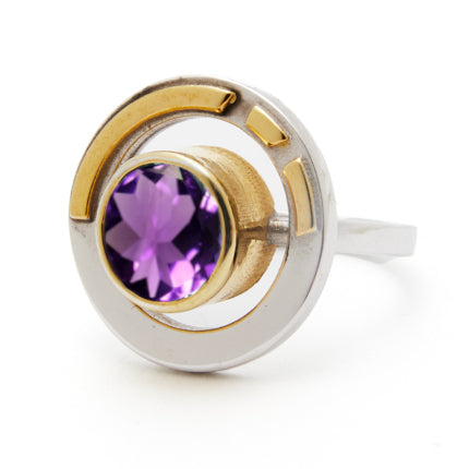 The Elodie Ring in Silver Gold & various gemstones by Gallardo & Blaine Designs showcases a round silver band adorned with a gold accent and a central purple gemstone. Blending vintage style glamour with its intricate, modern design, circular elements bea
