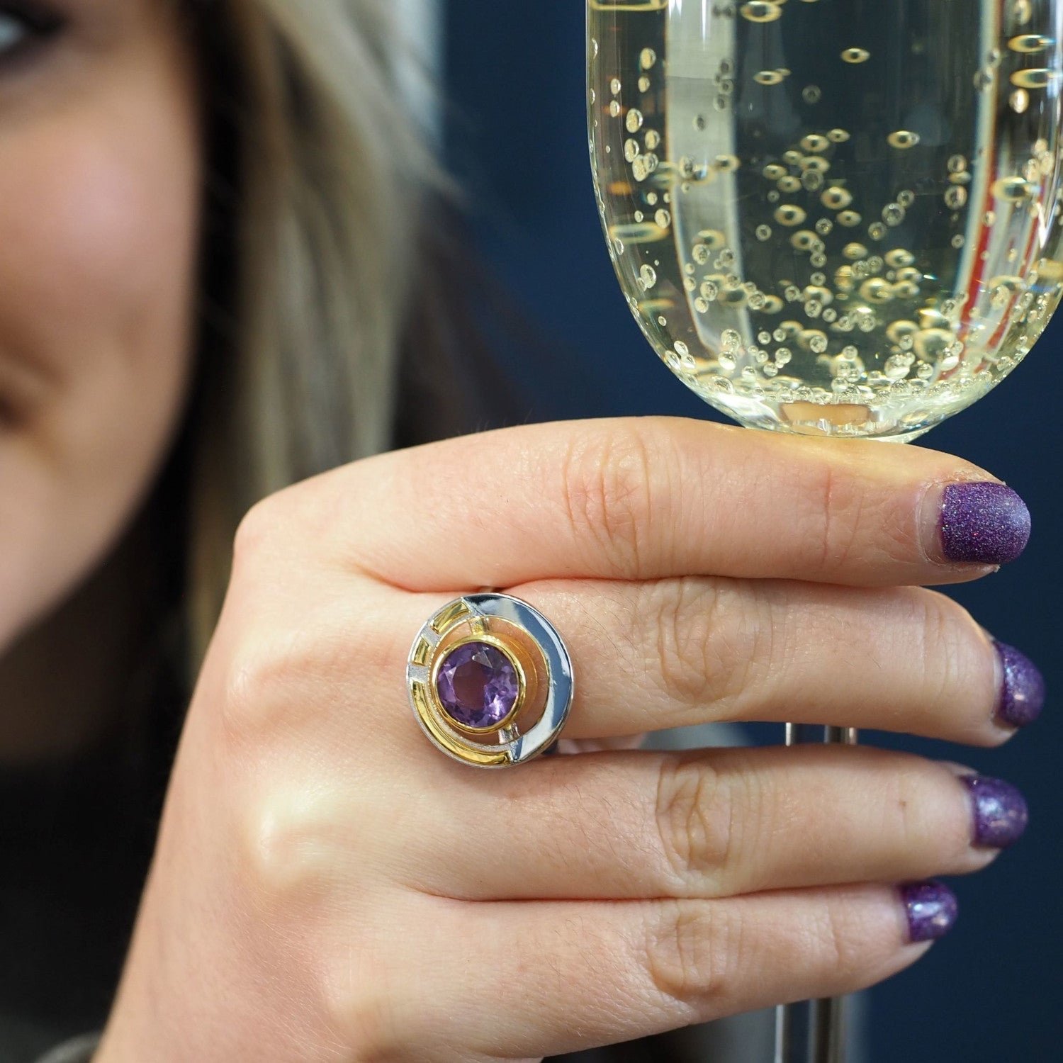 A person with purple glitter nail polish holds a glass of bubbly champagne. They are wearing the Gallardo & Blaine Designs Elodie Ring in Silver Gold & various gemstones, which features a large purple gemstone set in a gold and silver design. Part of thei