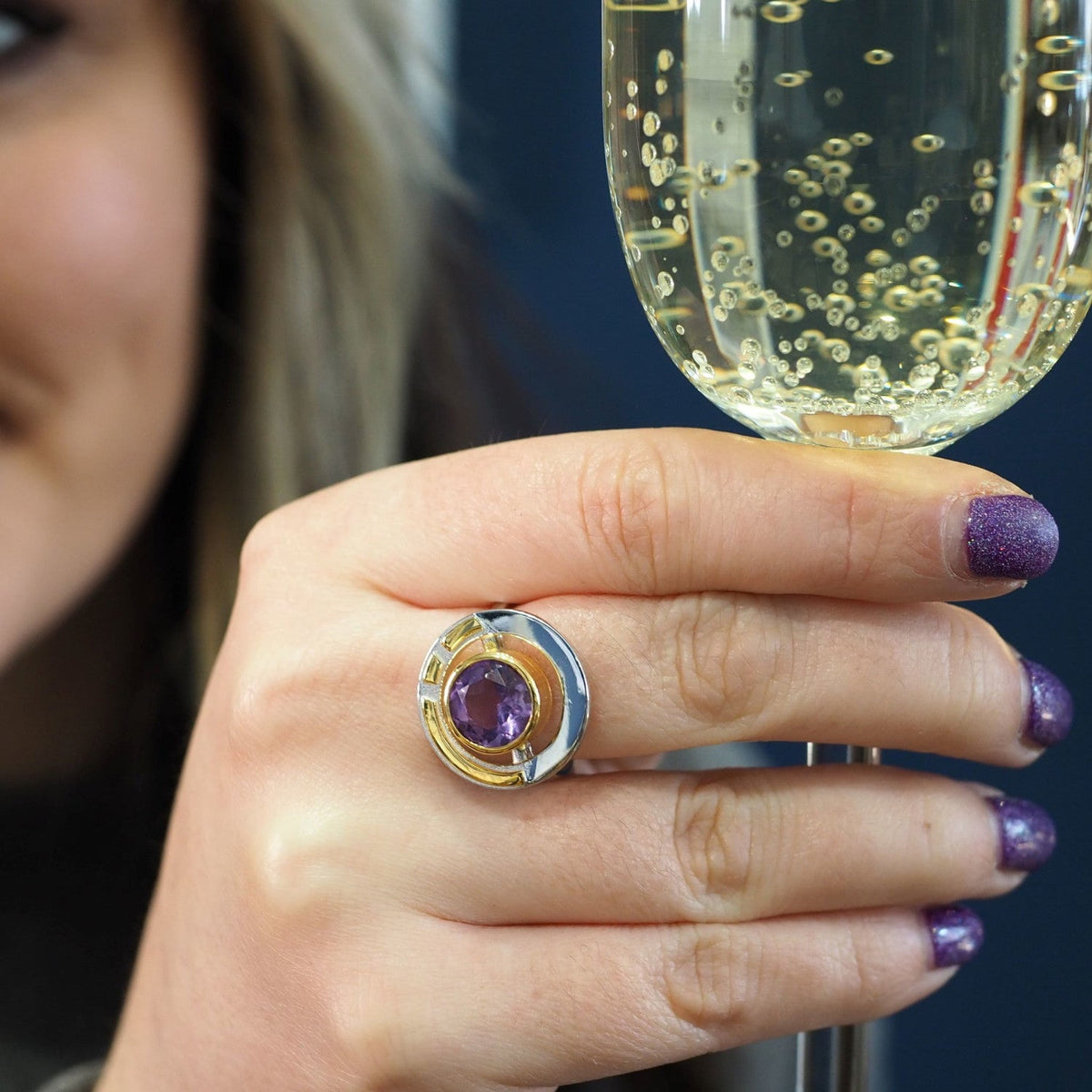 A person with purple glitter nail polish holds a glass of bubbly champagne. They are wearing the Gallardo & Blaine Designs Elodie Ring in Silver Gold & various gemstones, which features a large purple gemstone set in a gold and silver design. Part of thei