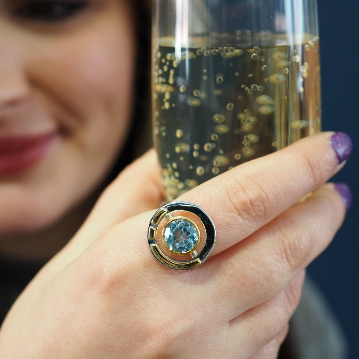 Close-up of a person holding a glass of sparkling beverage. The focus is on their hand, adorned with the Gallardo & Blaine Designs Elodie Ring in Silver Gold & various gemstones, exuding vintage style glamour. The person's face is slightly blurred in the 