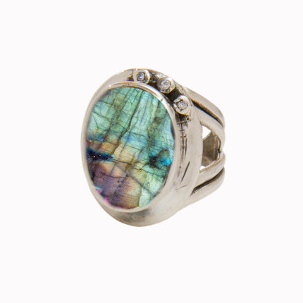 A silver statement cocktail ring featuring an oval-shaped labradorite stone with a spectrum of iridescent colors. The stone is set in a bezel setting, and the ring design includes three small round accent stones on one side of the band, enhanced by colour