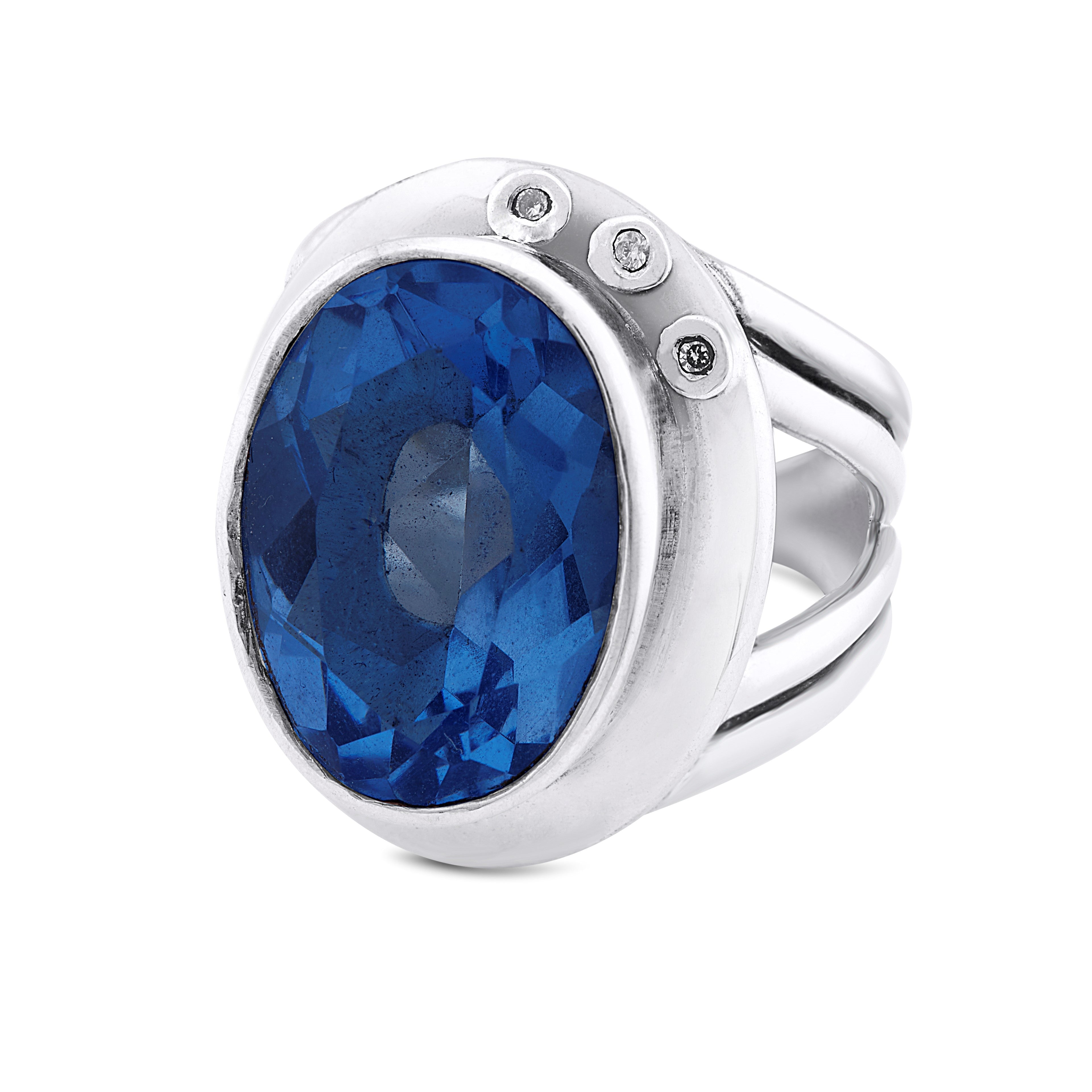 A stunning statement cocktail ring, the Eyetelia Ring in various gemstones by Gallardo & Blaine Designs features a large oval blue gemstone at its center, flanked by three small round white gemstones on one side. The band has a split shank style and an ad