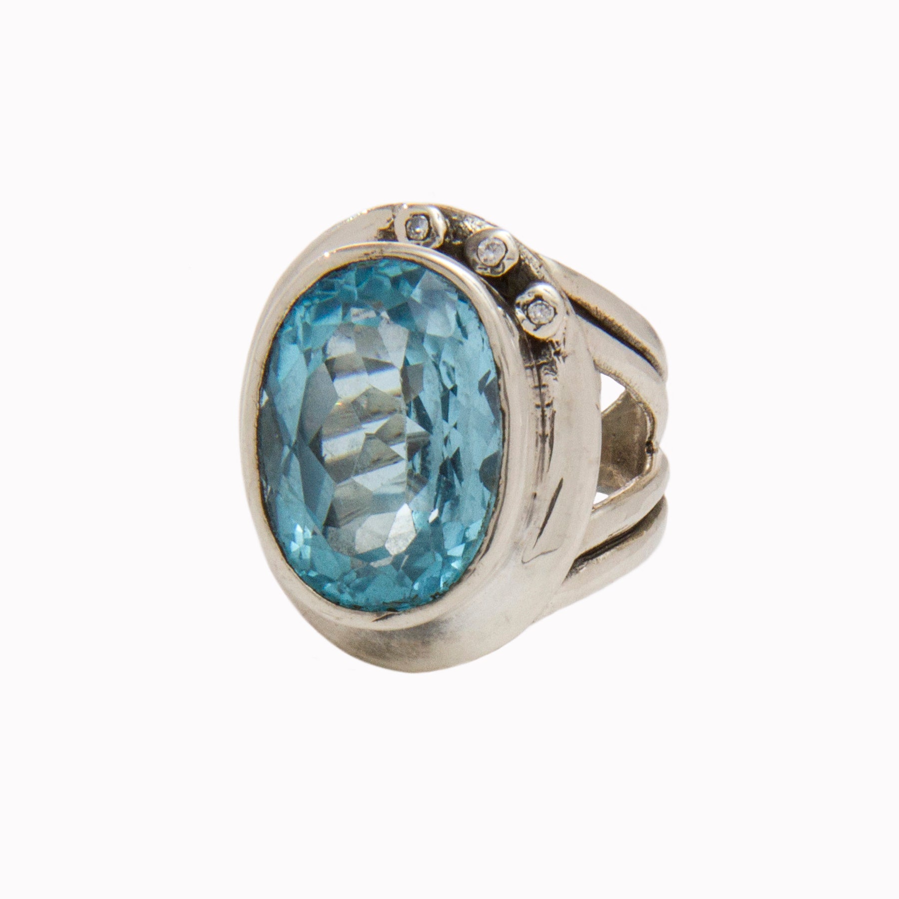 The Eyetelia Ring in various gemstones by Gallardo & Blaine Designs features a large oval-shaped blue gemstone set in the center. The faceted gemstone enhances its sparkle, while the band is adorned with three small round diamonds on one side, making it p