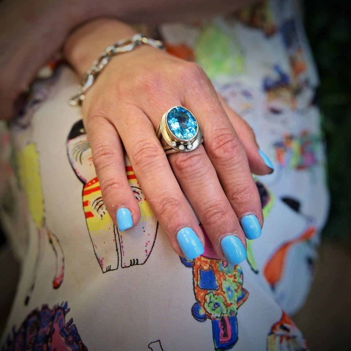 A hand with light blue nails wears the Gallardo & Blaine Designs Eyetelia Ring in blue gemstone, paired with a silver bracelet, resting on colorful cat-patterned fabric.