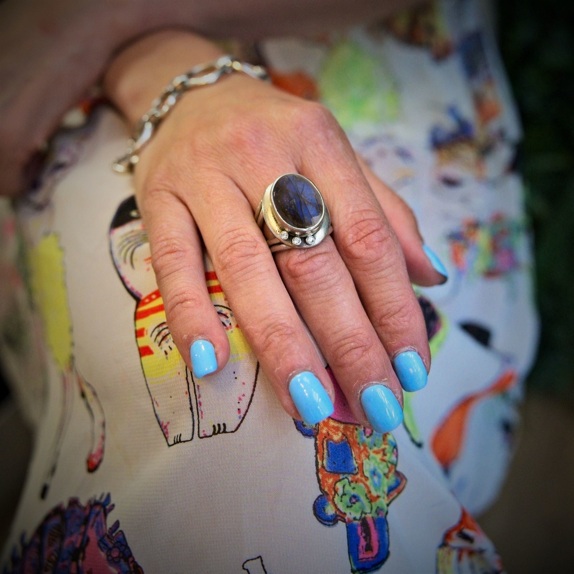 A hand with light blue nail polish wears the Gallardo & Blaine Designs Eyetelia Ring in a gemstone and a sterling silver gold vermeil bracelet, resting on colorful fabric with cartoon cats.