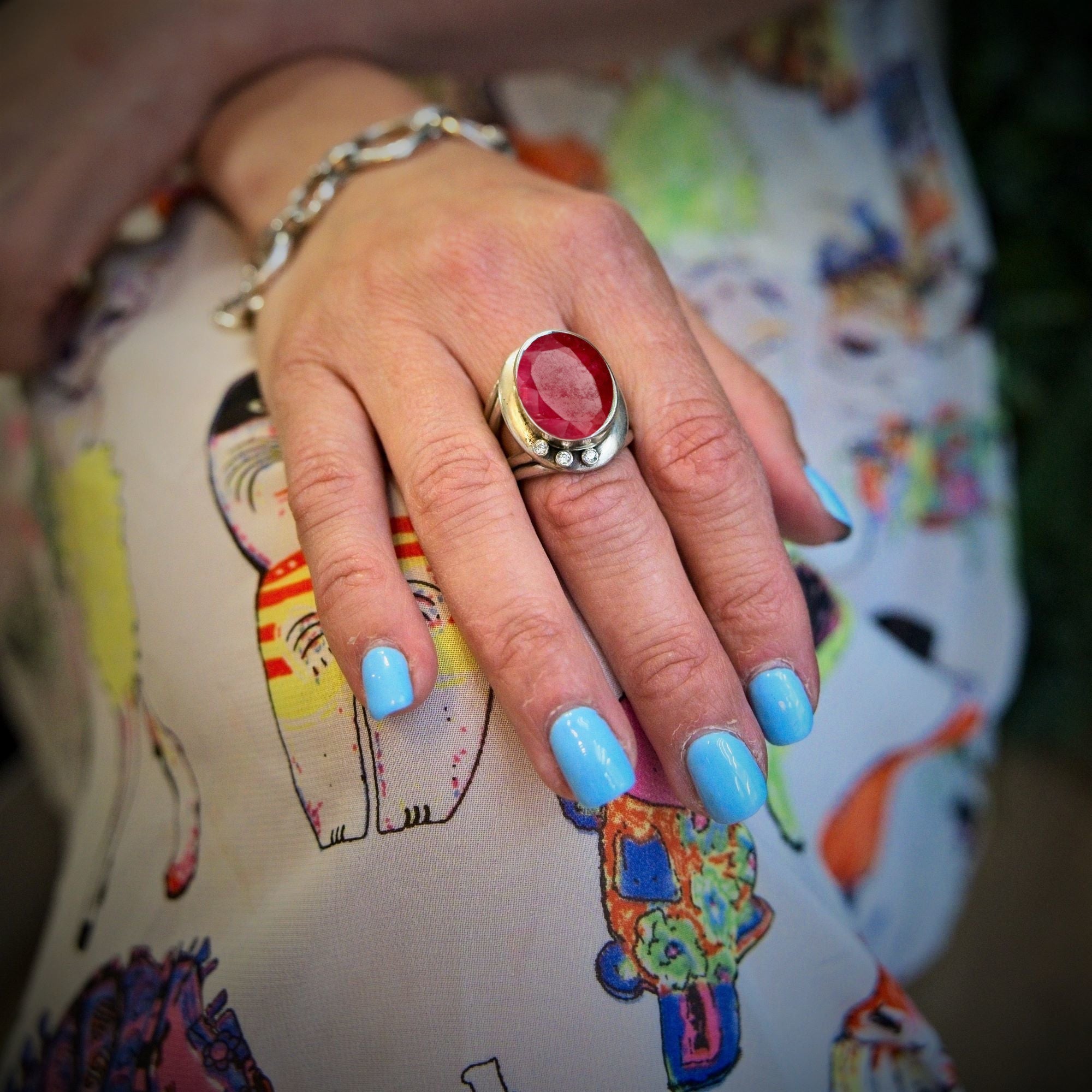 A hand with light blue polish wears the Eyetelia Ring by Gallardo & Blaine Designs—a bold silver band set with a red gemstone—resting on colorful cat-patterned fabric. A delicate silver bracelet adorns the wrist.