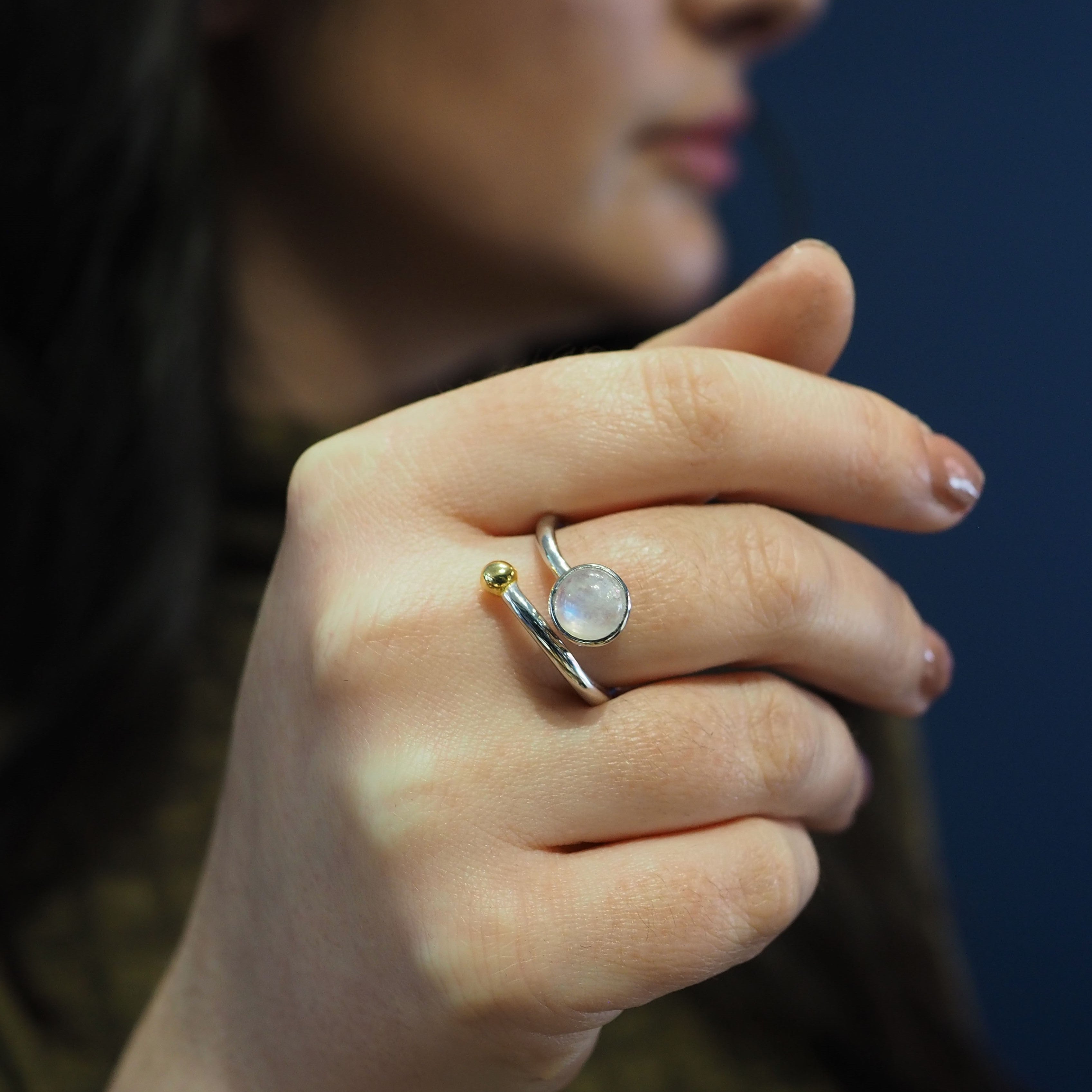Close-up of a person holding their hand near their face, showcasing an elegant adjustable Honeysuckle Ring in Silver Gold & gemstones. The silver and gold jewelry from Gallardo & Blaine Designs features a round, translucent stone set in a simple band desi