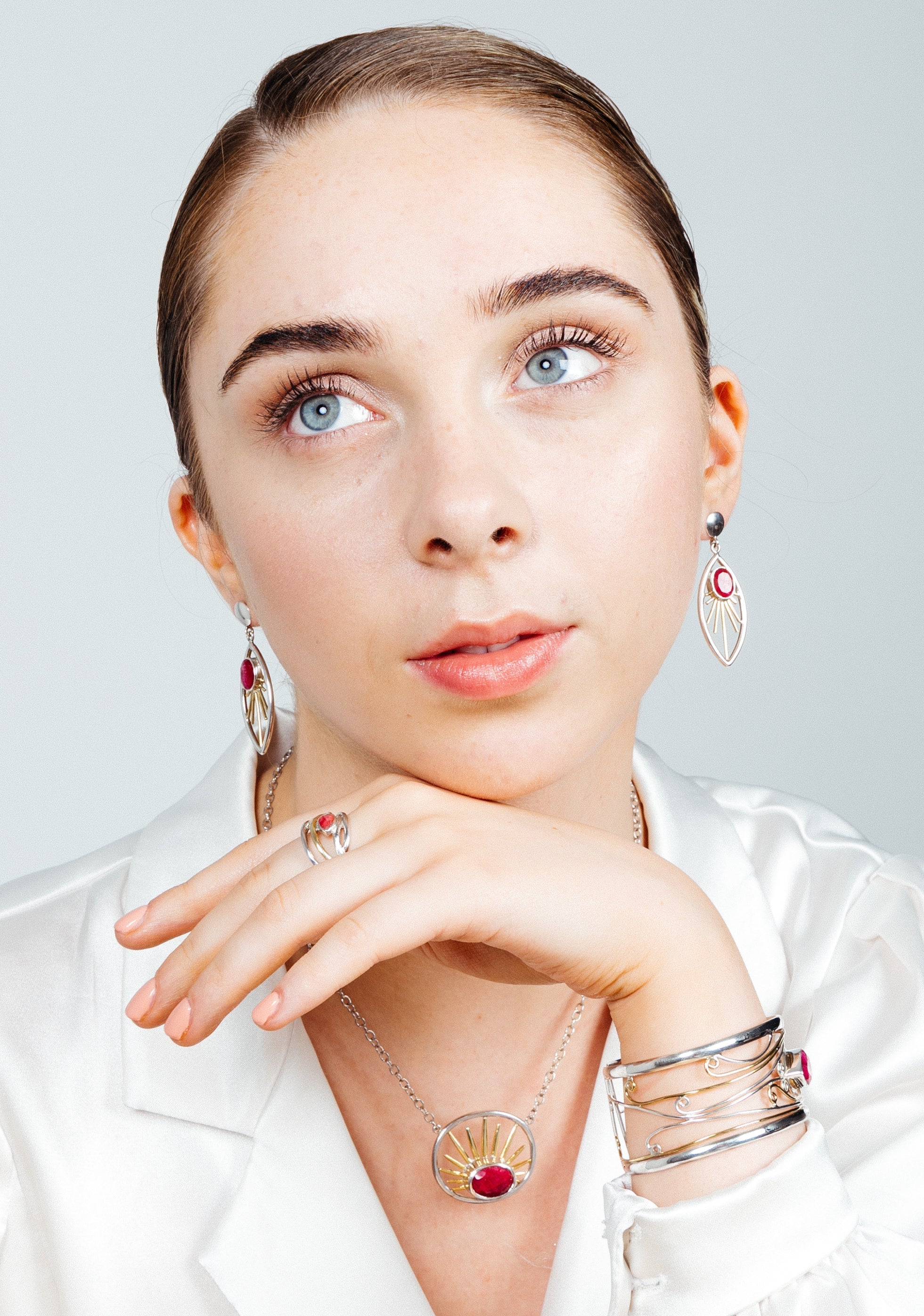 A young woman with light skin and light blue eyes is wearing a white blouse. She is adorned with Gallardo & Blaine Designs sterling silver jewelry, including a Goddess Necklace Small in various gemstones, earrings, a bracelet, and a ring, all featuring re