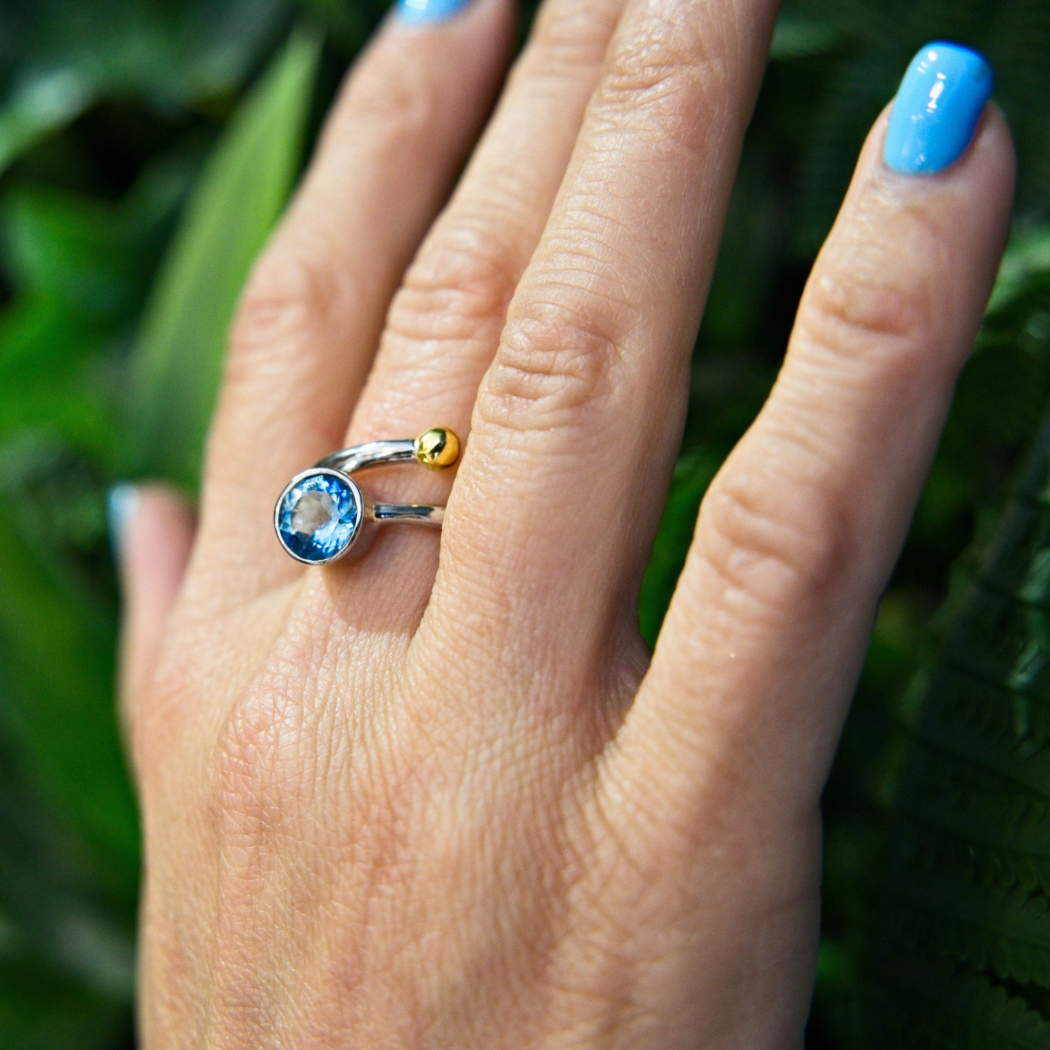A hand with blue nail polish wears an elegant Gallardo & Blaine Designs Honeysuckle Ring in Silver Gold & gemstones, showcasing a large, round blue gemstone and a small yellow bead. The silver ring, part of a stunning silver and gold jewelry collection, i