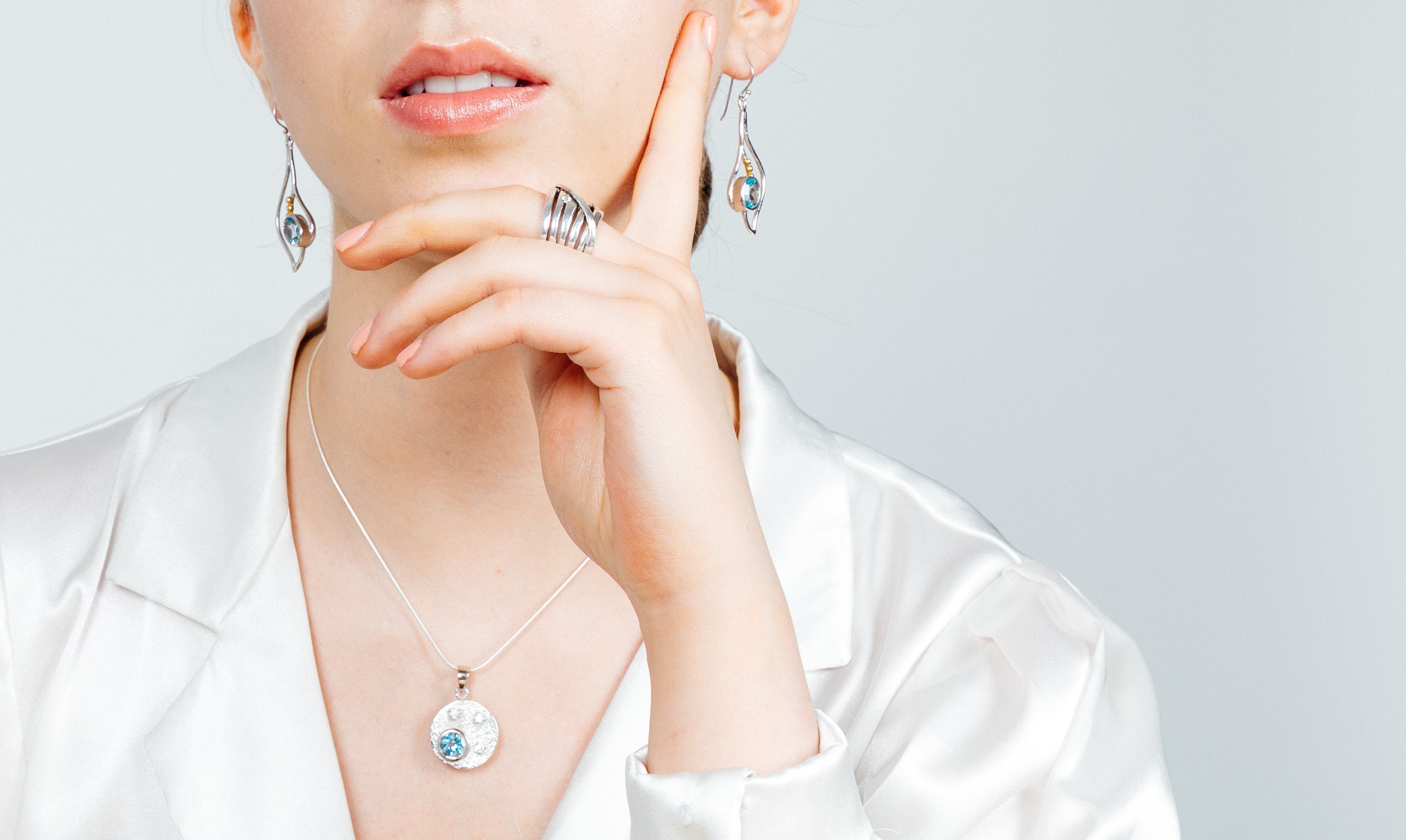 A person wearing elegant silver & gold jewelry, including Gallardo & Blaine Designs Iris Earrings in various gemstones, rings, and a necklace. The necklace features a round pendant with a blue gem. The person is dressed in a white satin blouse, with their