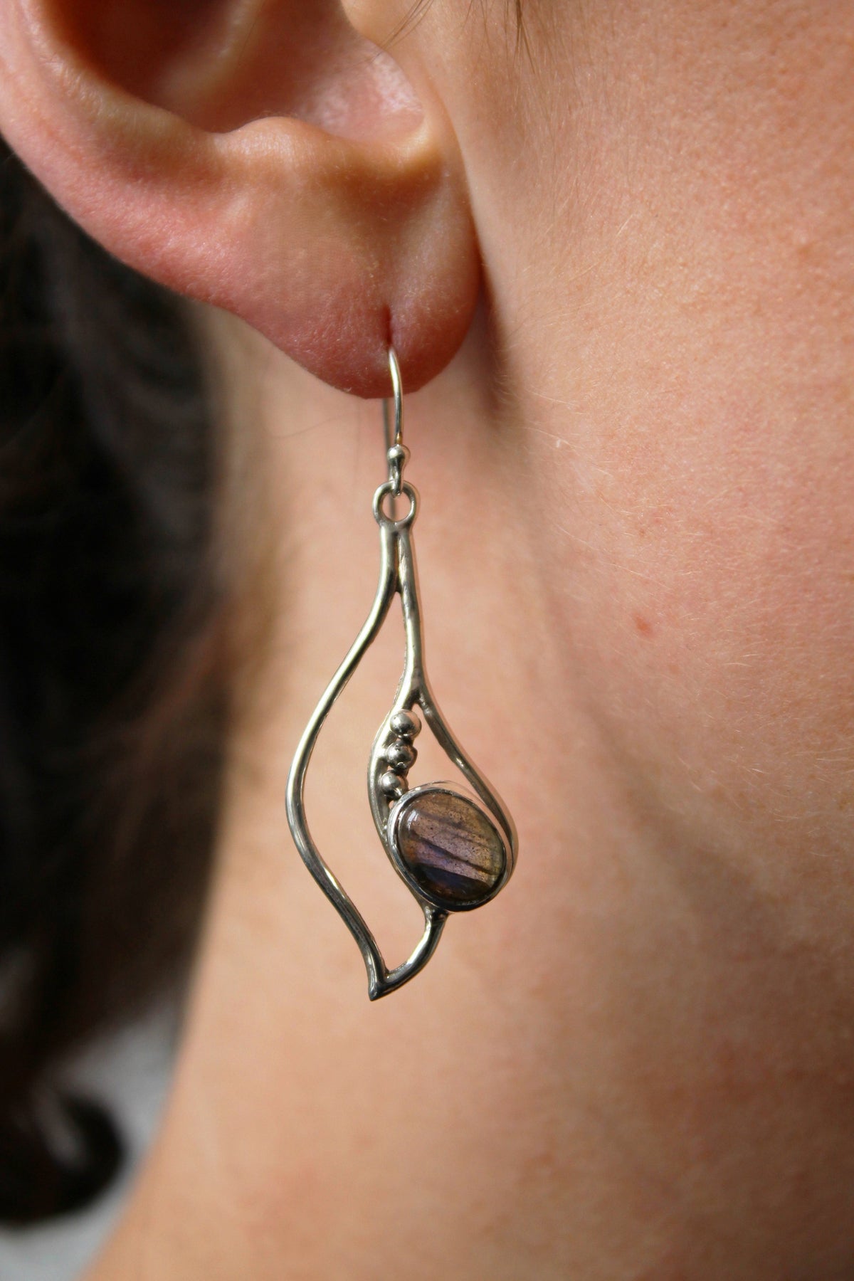 Close-up of a person's ear wearing Gallardo & Blaine Designs Iris Earrings—dangle earrings in polished silver & gold with an abstract shape and shimmering gemstone accents.