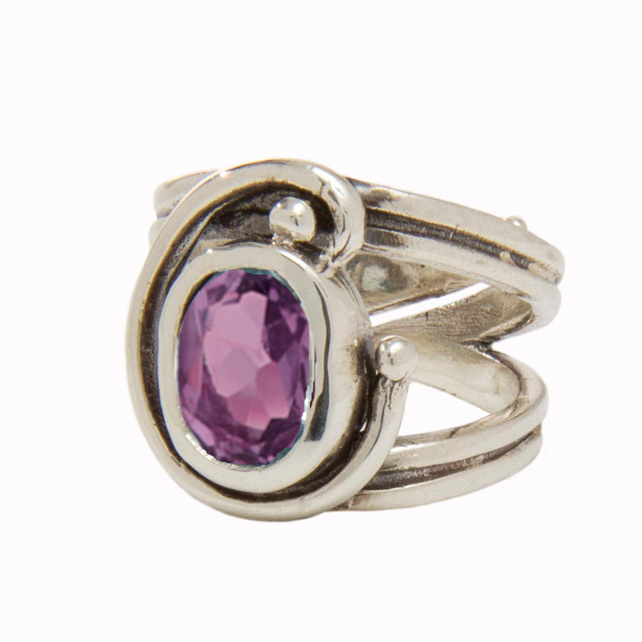 A silver Jasmine Ring in various gemstones by Gallardo & Blaine Designs features an oval-shaped, faceted purple gemstone set in the center, surrounded by an intricate swirl design. The thick silver band is split into three parts where the swirl design att