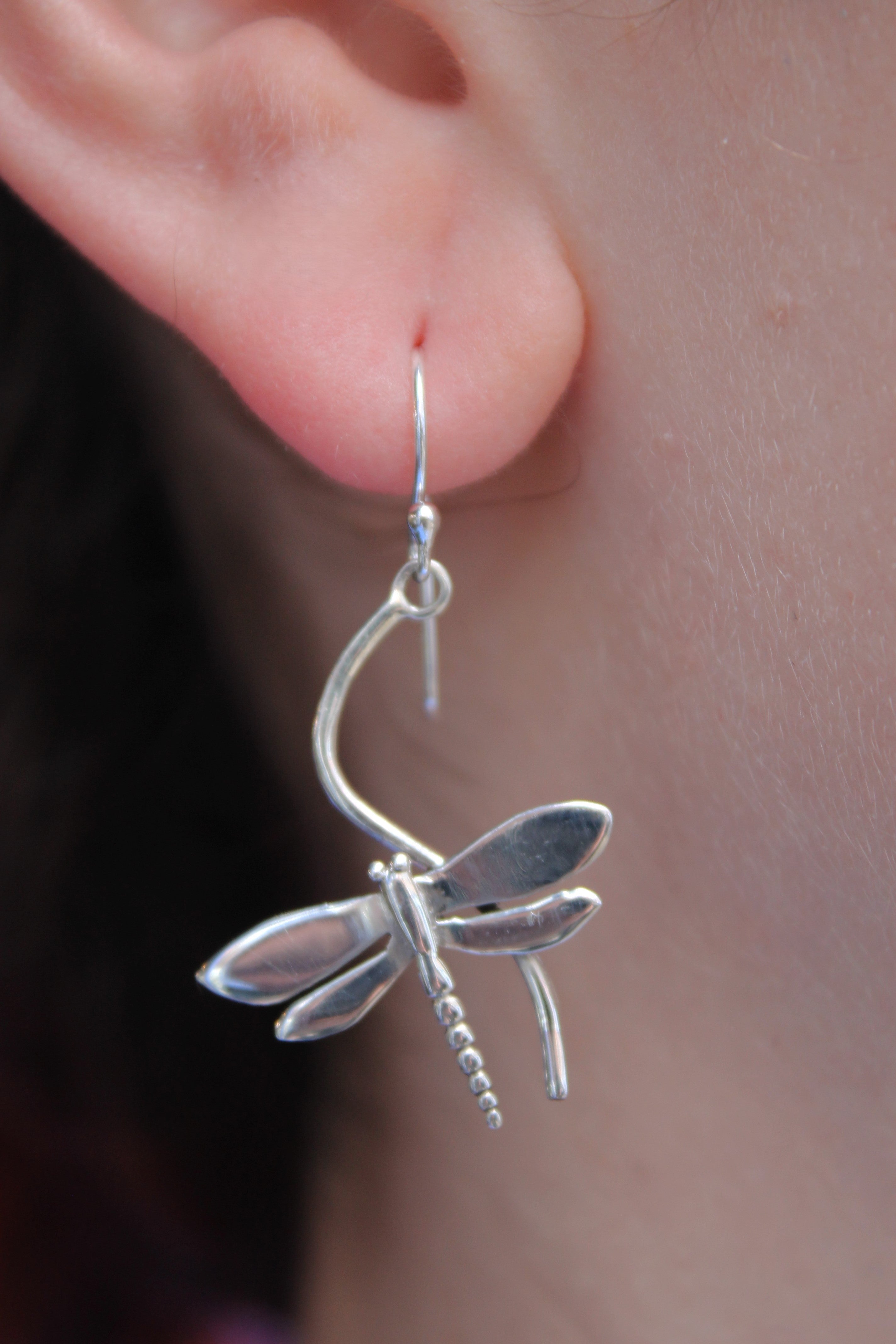 A close-up of a person's ear adorned with a detailed silver dragonfly from Watermelon Tropical's Wildlife Juniper Earrings, available in silver or gold and featuring lifelike wings, gracefully dangling from a simple hook.