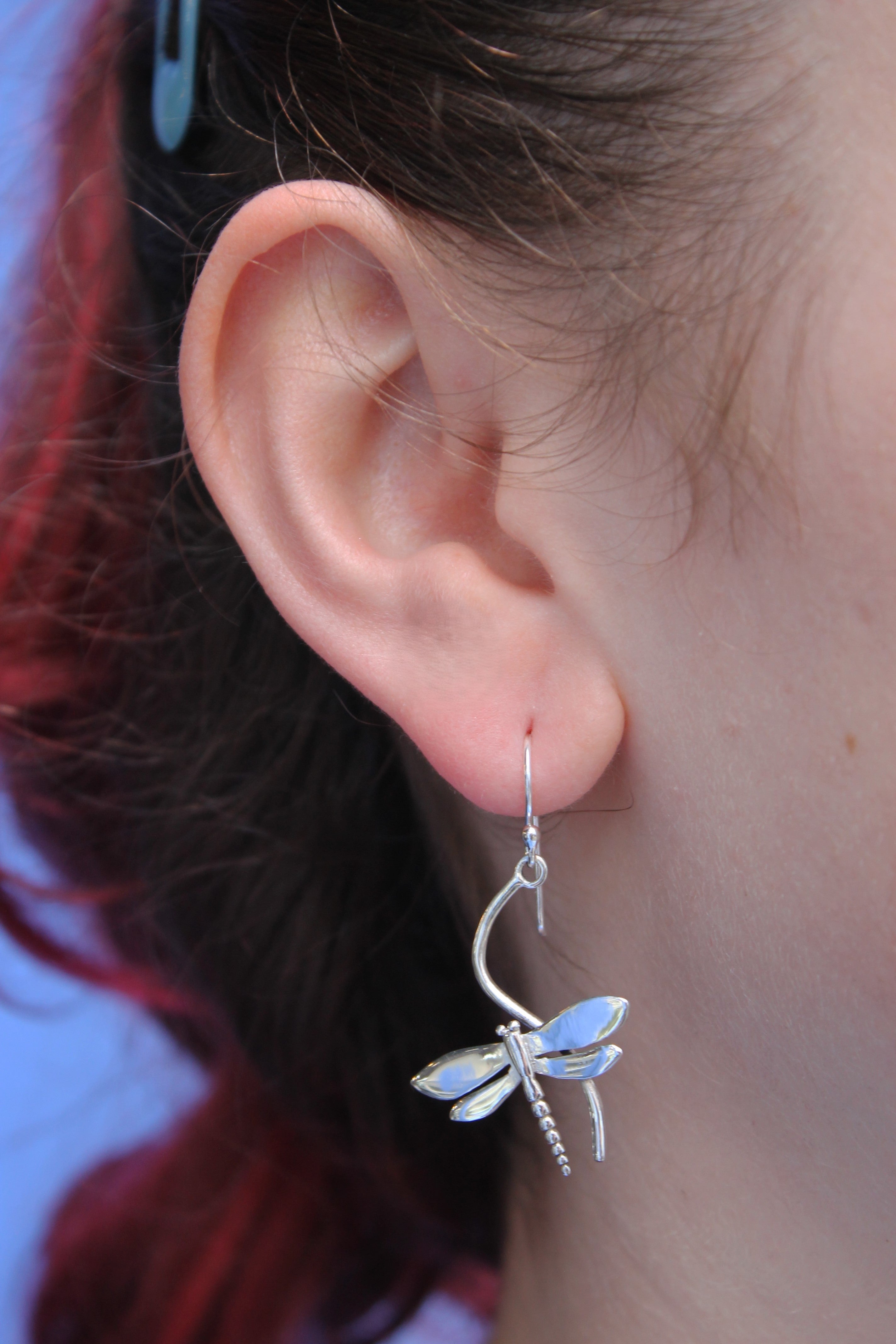 Close-up of a person's ear wearing Watermelon Tropical Wildlife Juniper Earrings (Silver or Gold), featuring iridescent dragonfly wings. The person has reddish hair pulled back with a blue clip, set against a light blue background.