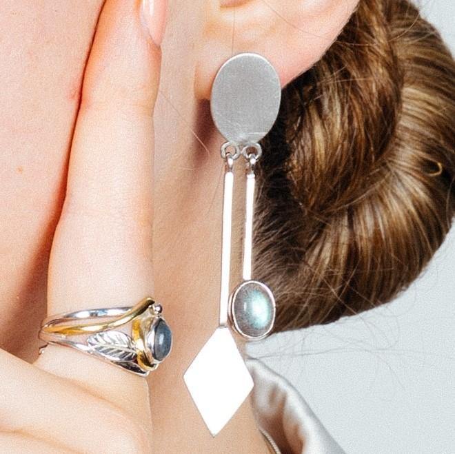 Close-up of a person's ear wearing the elegant Karo Earrings in various gemstones by Gallardo & Blaine Designs, featuring oval and diamond-shaped elements with a reflective surface. The person also wears a silver ring with a large, round, greyish-blue sto