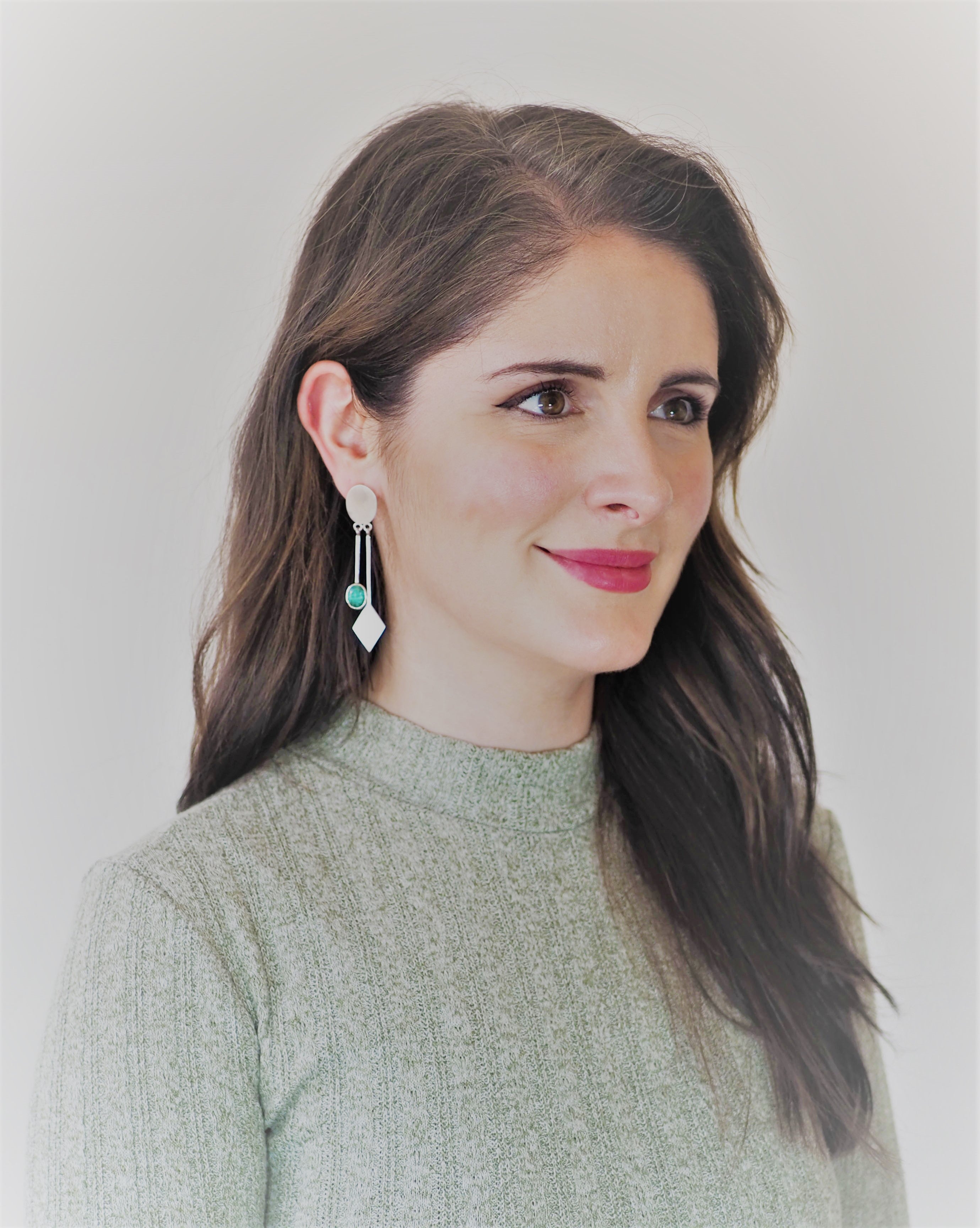 A woman with long brown hair and a light green textured top gazes to the right, highlighting Gallardo & Blaine Designs' Karo Earrings. These silver dangle drop earrings elegantly mix circular and diamond shapes against a softly blurred background.