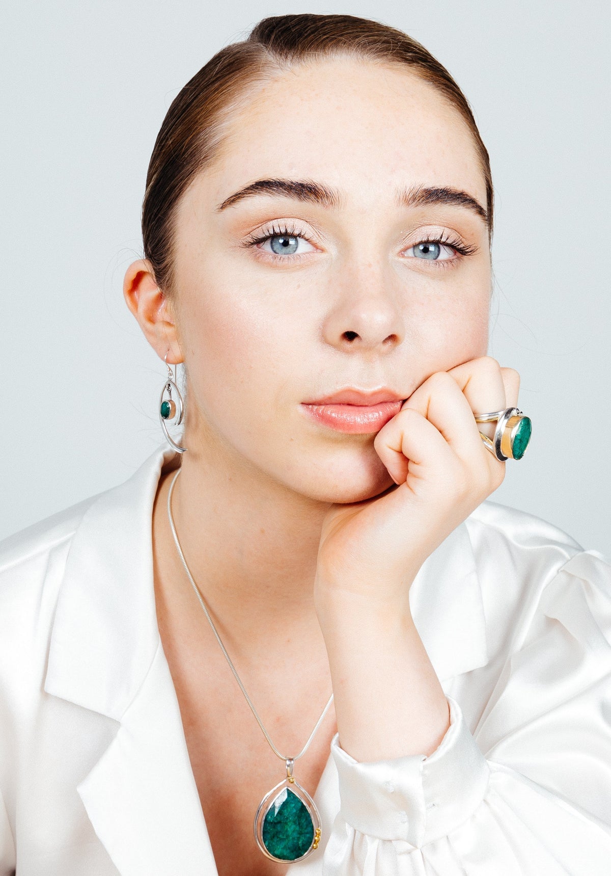 A woman with light skin and blue eyes gazes directly into the camera, resting her chin on her hand. She exudes minimalist elegance in a white blouse and matching green gemstone jewelry, including a necklace, Primrose Earrings in various gemstones from Gal