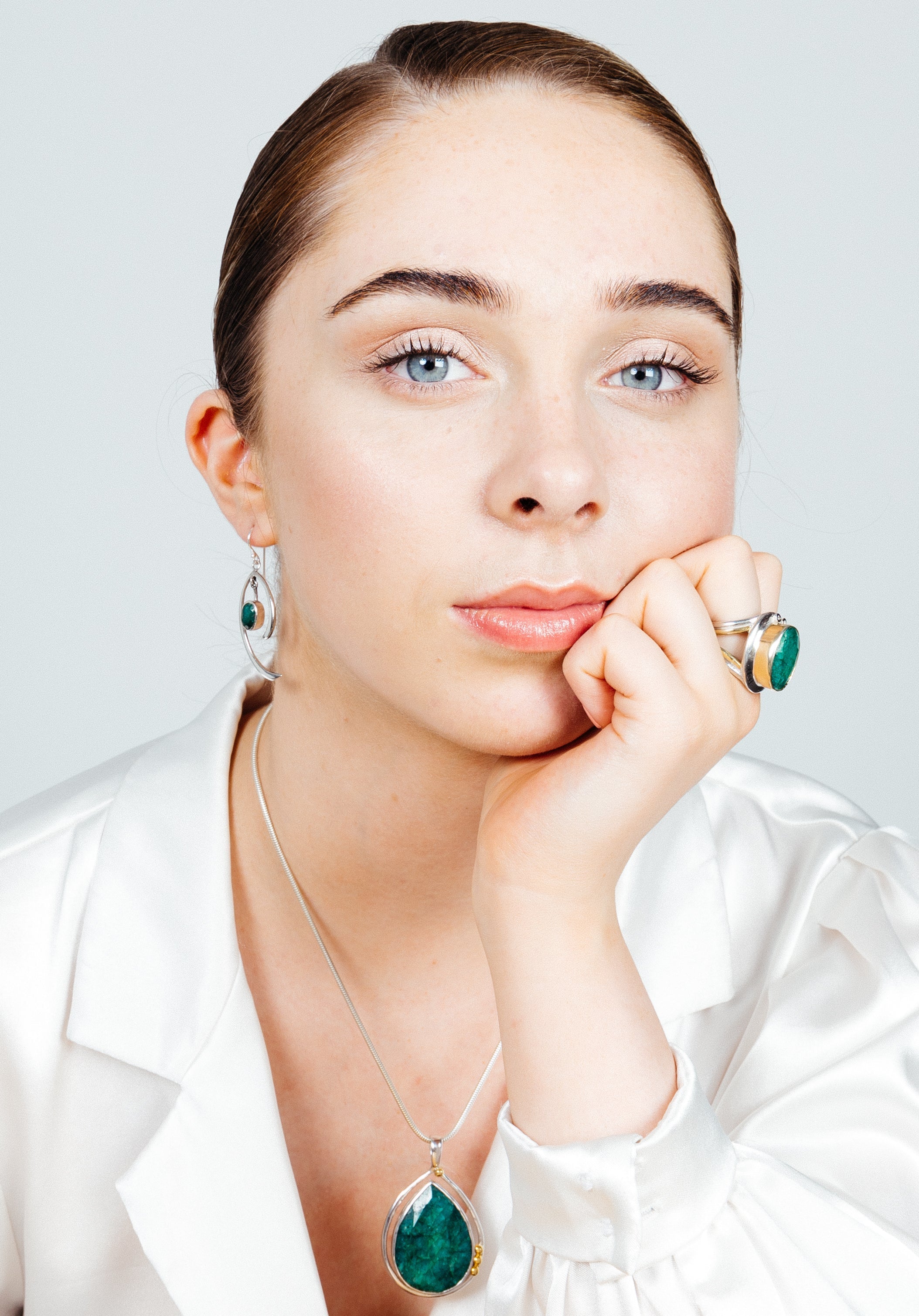 A woman with light skin and blue eyes gazes directly into the camera, resting her chin on her hand. She exudes minimalist elegance in a white blouse and matching green gemstone jewelry, including a necklace, Primrose Earrings in various gemstones from Gal
