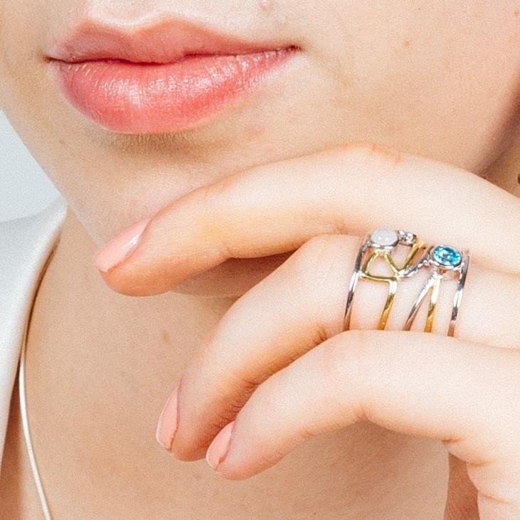 Close-up of a person’s face and hand. The person is wearing a Gallardo & Blaine Designs Lattice Ring in various gemstones—blue, clear, and gold accents—on their index finger. Their lips are softly closed, and the hand adorned with contemporary design touc