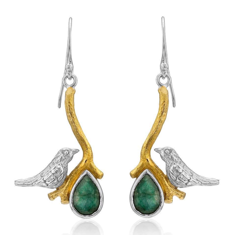 The Lovebird Earrings in various gemstones by Gallardo & Blaine Designs are a pair of elegant statement earrings featuring a small silver bird perched on a golden branch. Each branch extends down to hold a green teardrop-shaped gemstone. These dangle drop