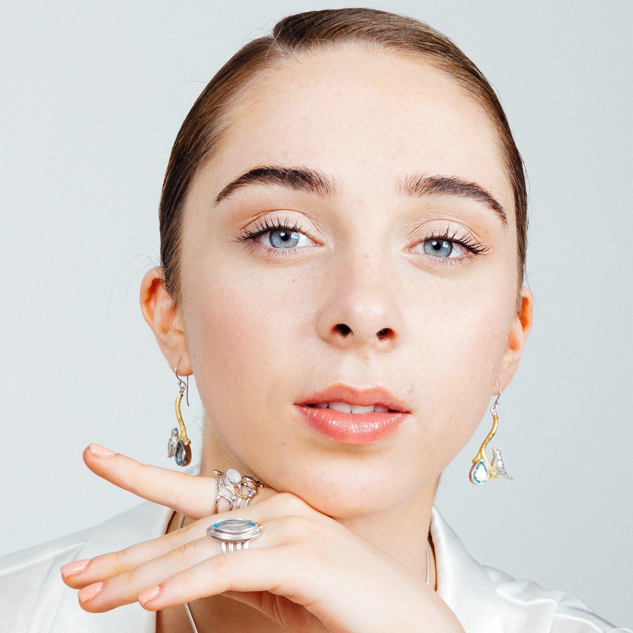 A young woman with fair skin and blue eyes gazes into the camera. She wears Gallardo & Blaine Designs' Lovebird Earrings in various gemstones and a ring with a large stone on her finger, which is gently resting near her chin. She has light makeup and her 