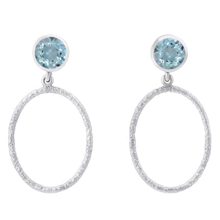 A pair of Gallardo & Blaine Designs Lunar Earrings in various gemstones featuring a small round blue gem at the top, attached to a large textured open circular hoop. The design is simple yet elegant, with the blue gems adding a touch of color to the sleek